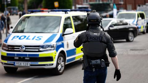 A police officer in tactical gear stands in front of a Swedish police car