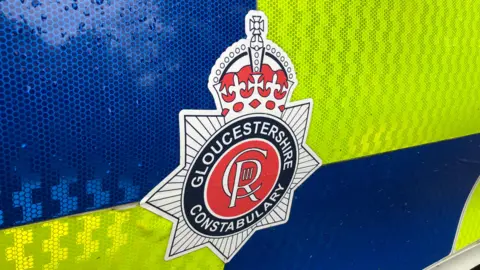 A close up of the Gloucestershire Constabulary badge on a police car door which has blue and yellow hi-vis chequering. The badge includes the "Charles Royal" emblem and a crown.