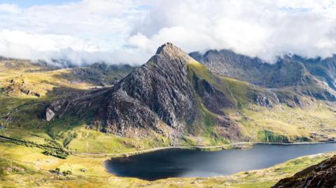 Landscape image showing Tryfan peak, with Ogwen lake below on a sunny day with low cloud behind the mountain covering other peaks in the Caerneddau range