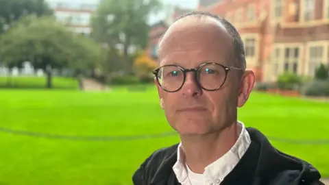 Prof Geraint Ellis standing in front of a lawn. He is unsmiling, wearing a white shirt and a hooded top and has horn-rimmed glasses