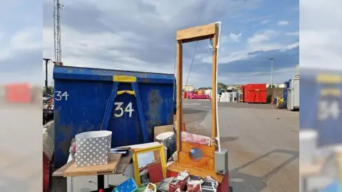 A wooden guillotine, with an electric motor and a blade splatted with fake blood, stands among a pile of rubbish at Grimsby waste recycling centre. A blue skip, for holding waste, stands to the left.