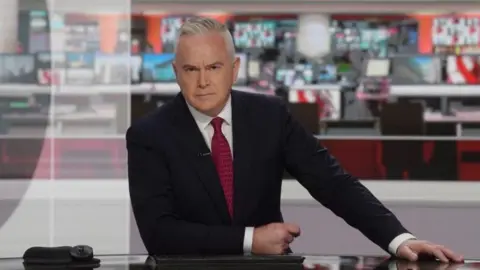 BBC Huw Edwards presenting the news in studio wearing suit and red tie