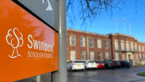 An orange Swindon Borough Council sign with council offices blurred in the background