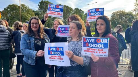 Five women outside school gates holding pieces of paper say 'Save Our School'