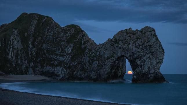 Durdle Door cliff arch in near darkness with the orange moon on the horizon in the centre of the arch