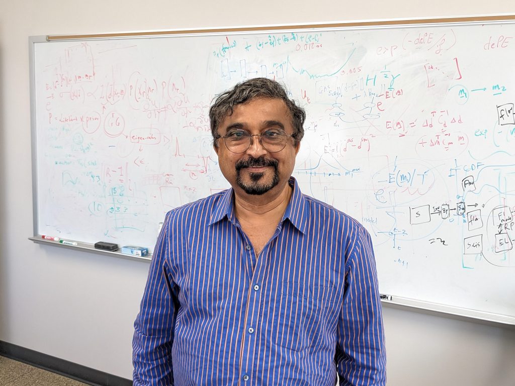 Photo of Mrinal in his office in front of a strategically placed whiteboard with equations.