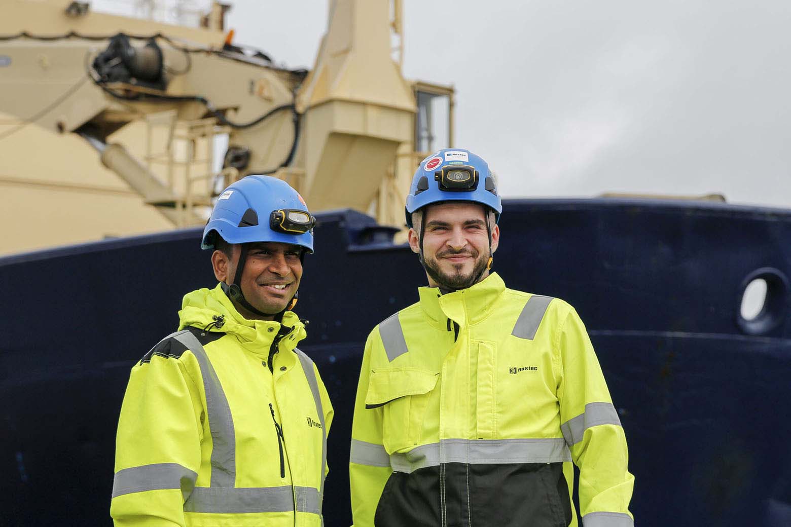 Two happy smiling men with blue helmets and neon yellow jackets with an icebreaker in the background