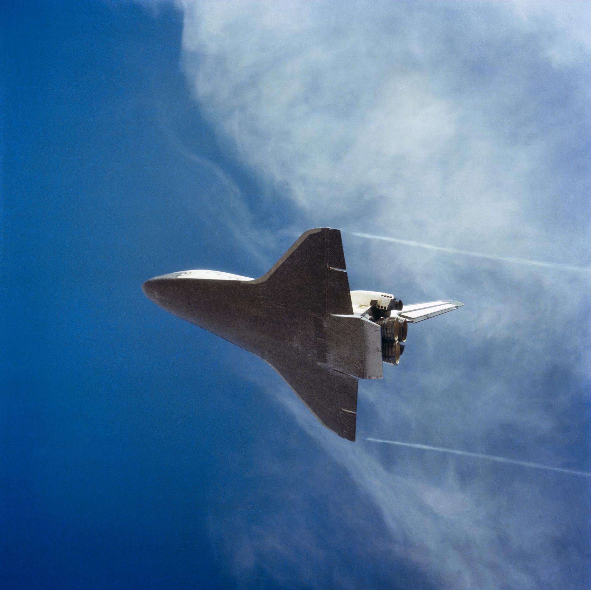 Space Shuttle Columbia (STS-2) is seen from below just before landing with the blue sky and wispy clouds visible in the background.