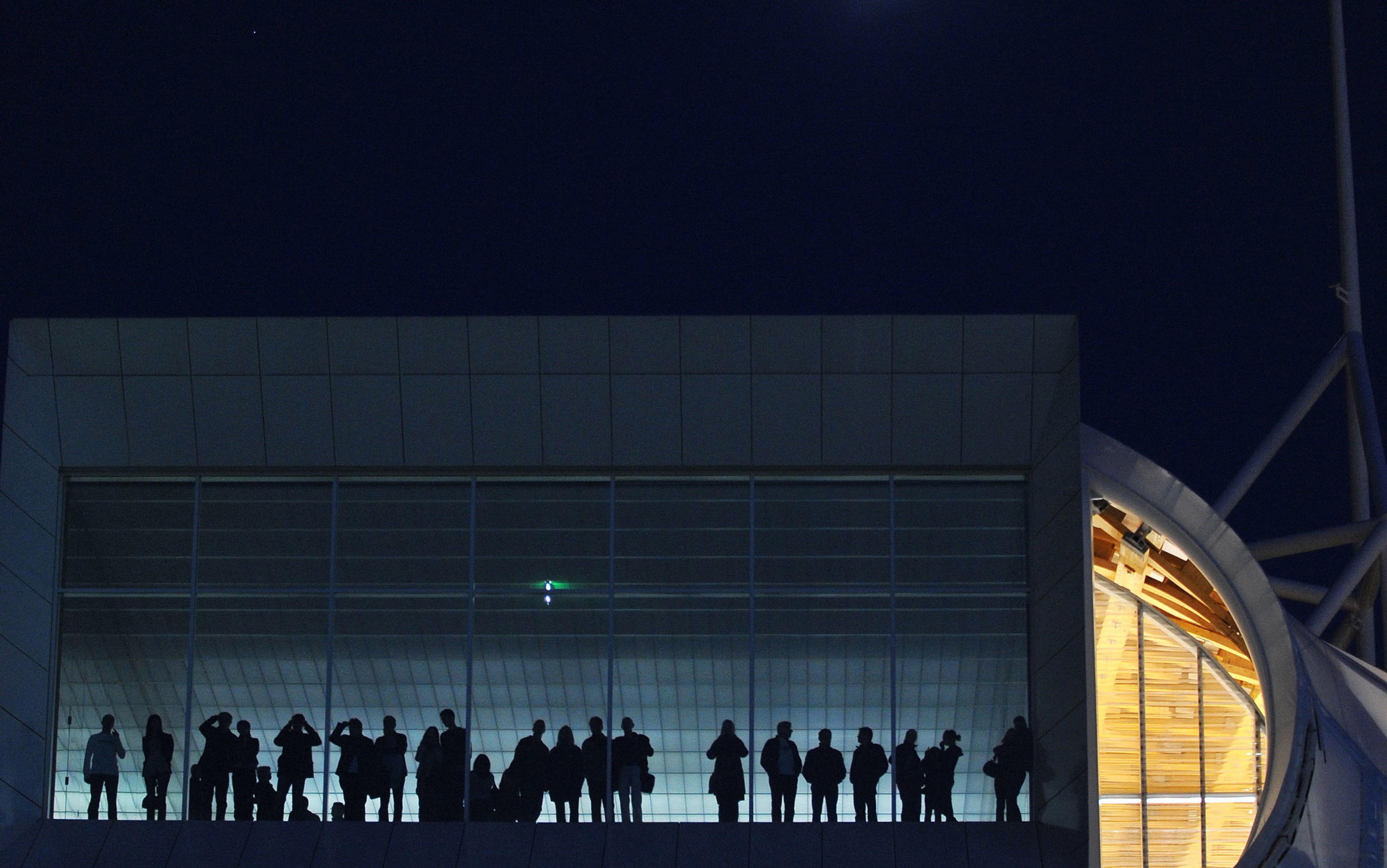 Silhouetted people stand inside a modern building with large windows and geometric architecture at night.