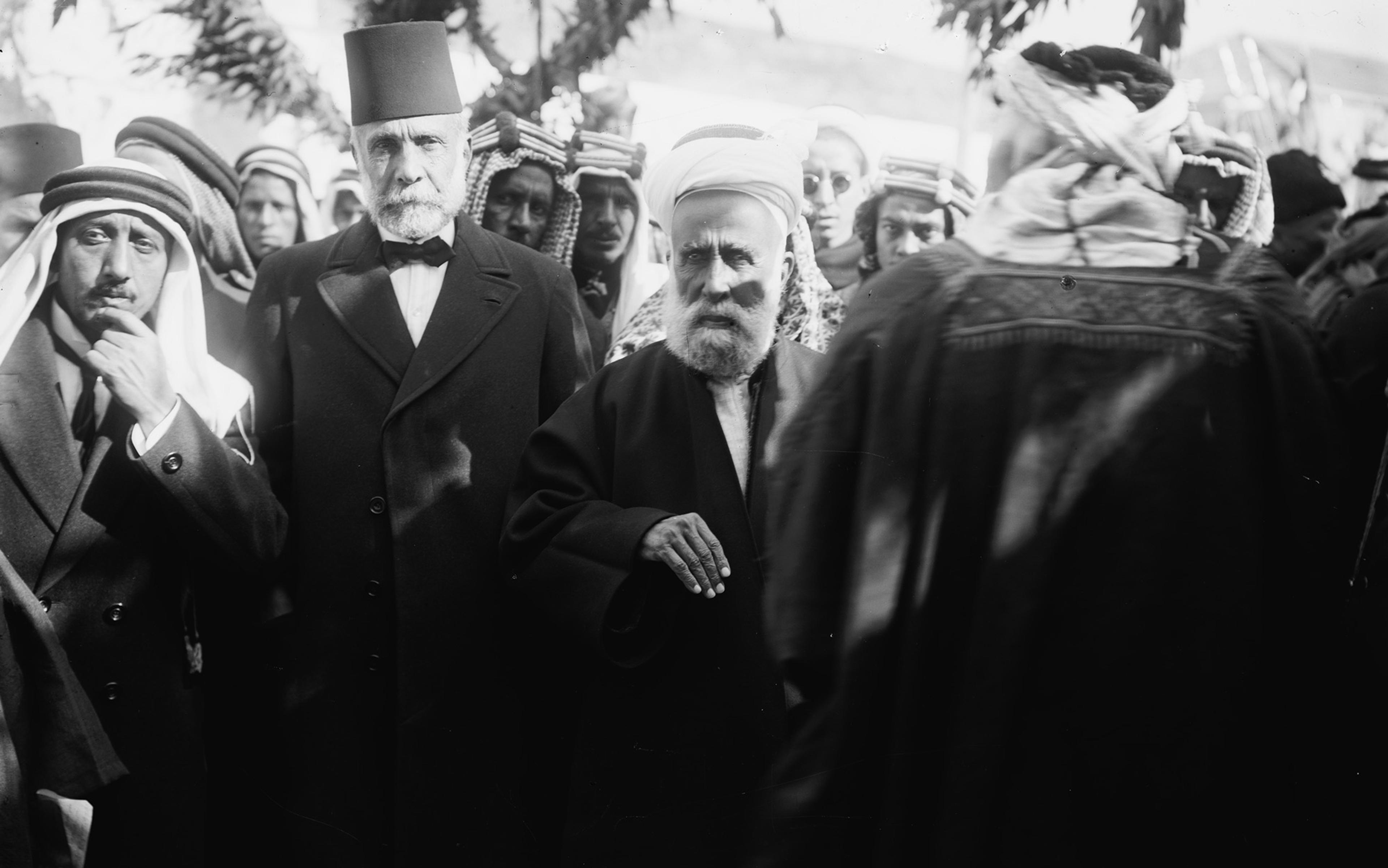 A group of men wearing traditional Middle Eastern attire and formal coats gathered outdoors, one man in focus with a fez.