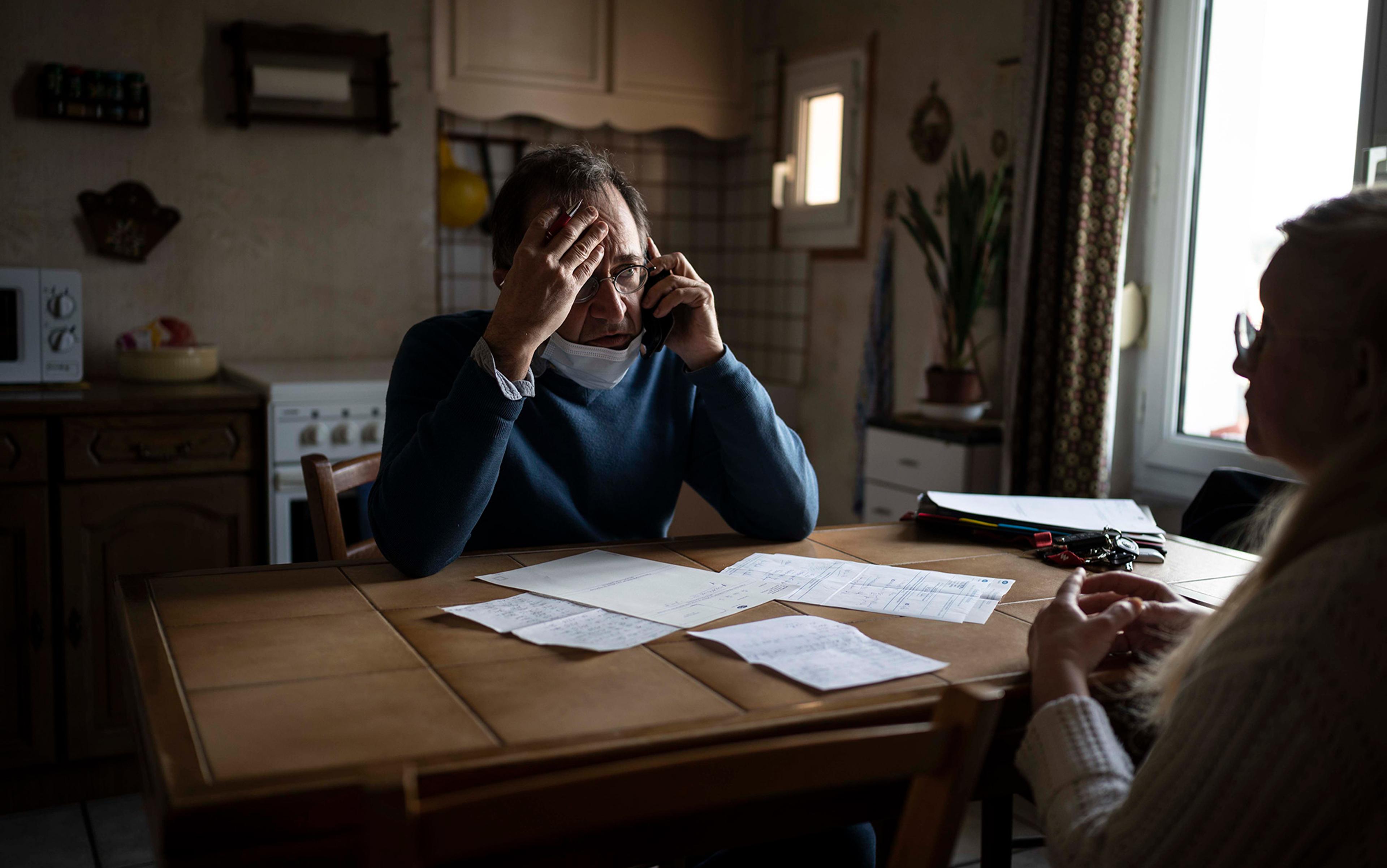 Man in a blue jumper wearing a surgical face mask on the phone, looking stressed at table with documents; woman with glasses sitting opposite him at home.