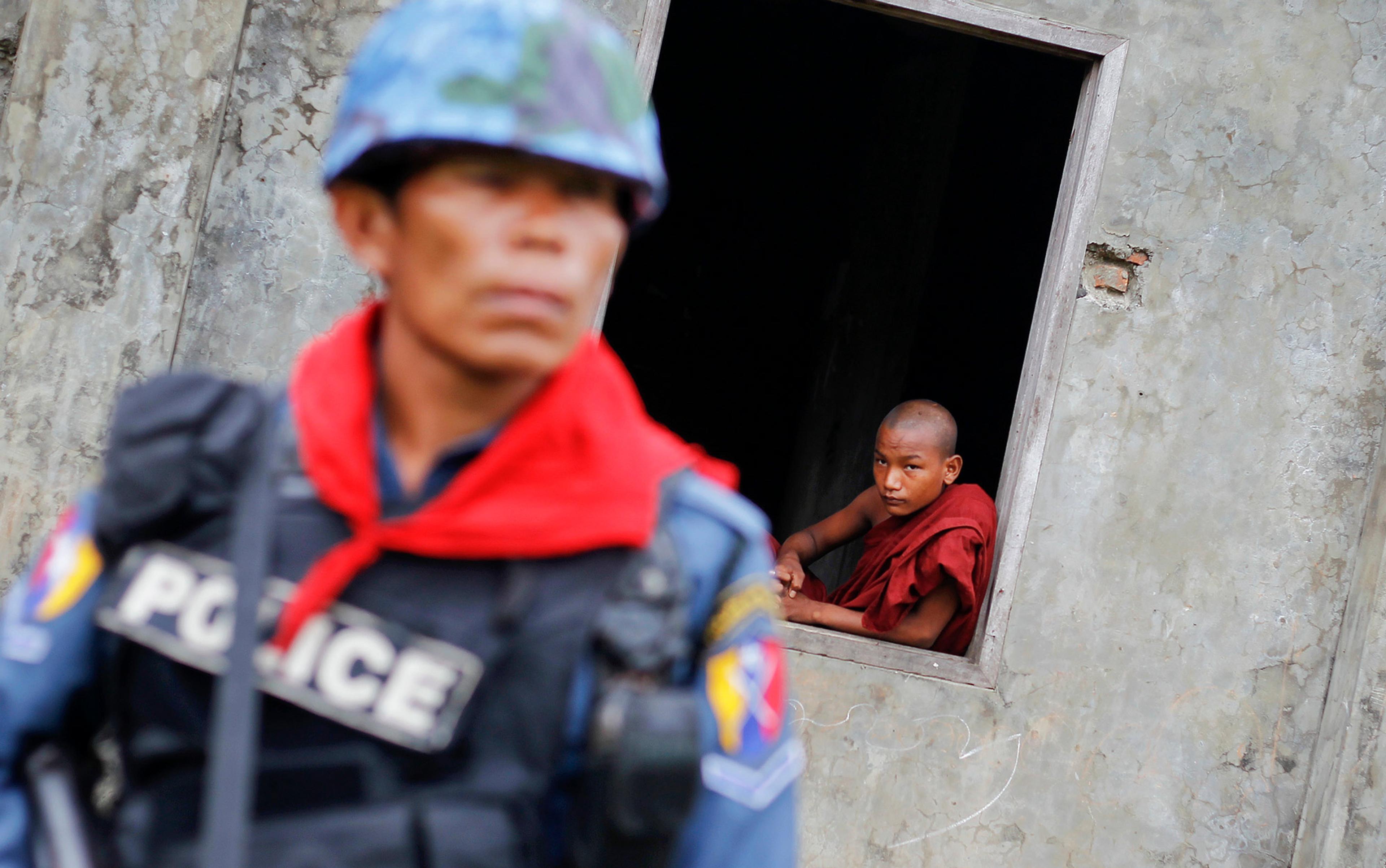 A uniformed police officer stands in the foreground, while a young monk in red robes looks out from a window of a concrete building in the background.