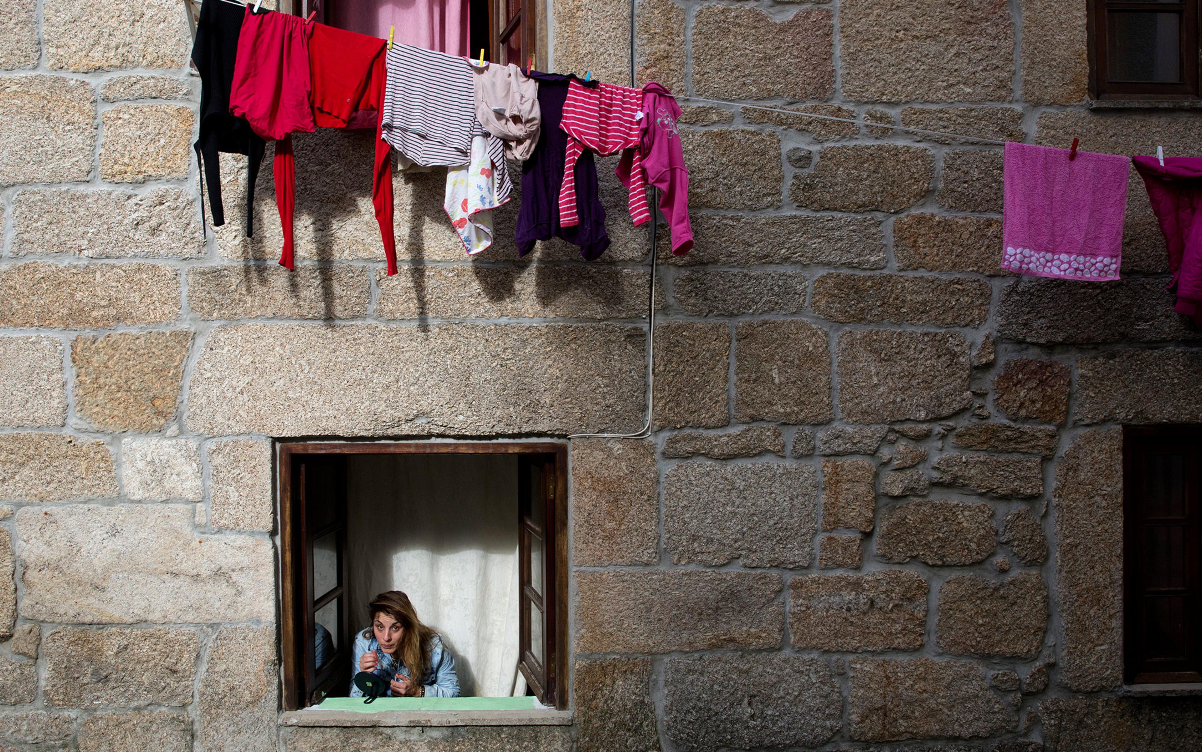 Woman looking out of a stone building window; clothes drying on a line extend from an upper window above her.