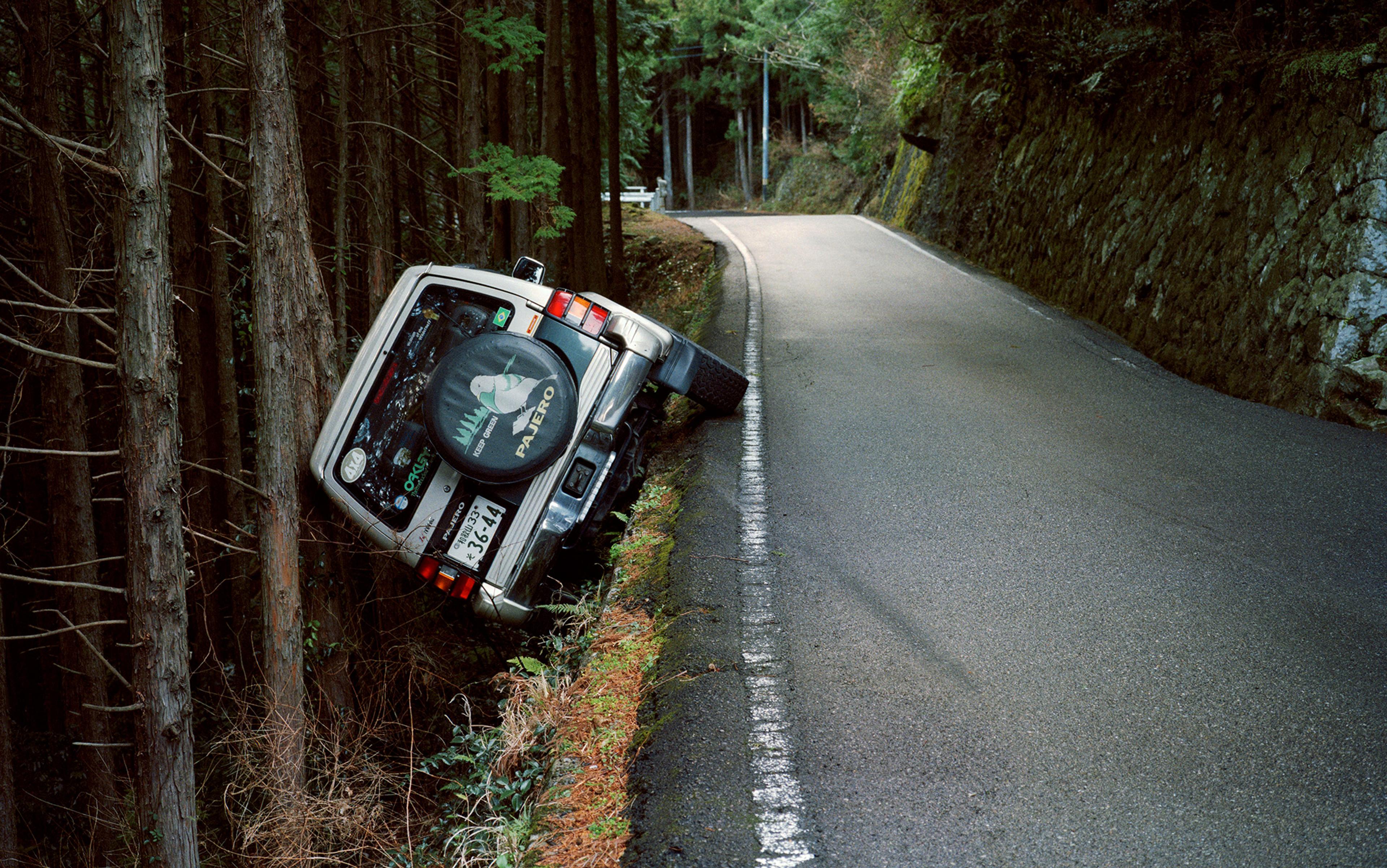 A narrow road in a forest with a white SUV that has veered off and lies on its side by the trees on the left side of the road.