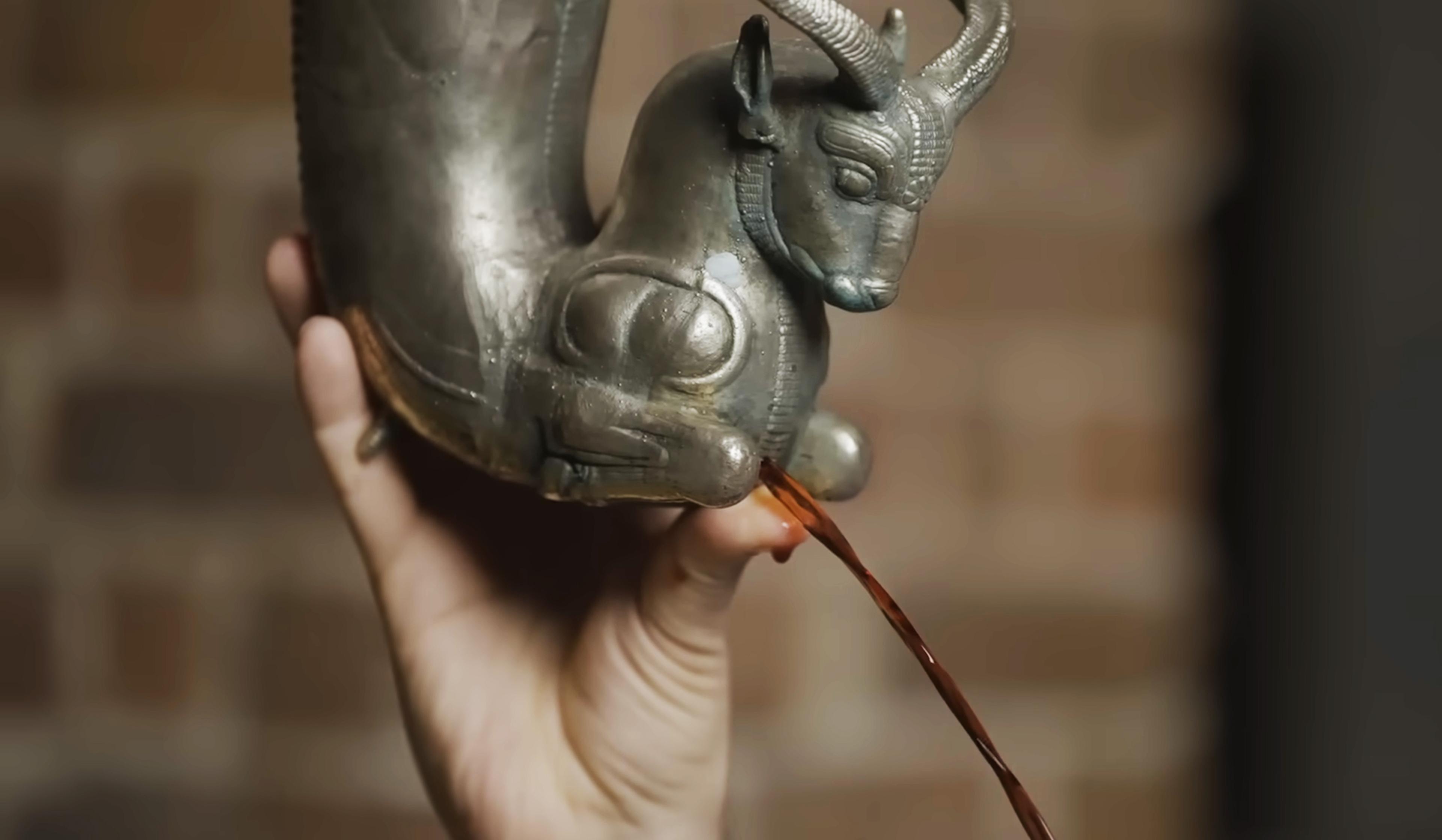 A hand holding an ancient horn-shaped bronze vessel with a sculpted animal head, pouring liquid against a brick wall background.