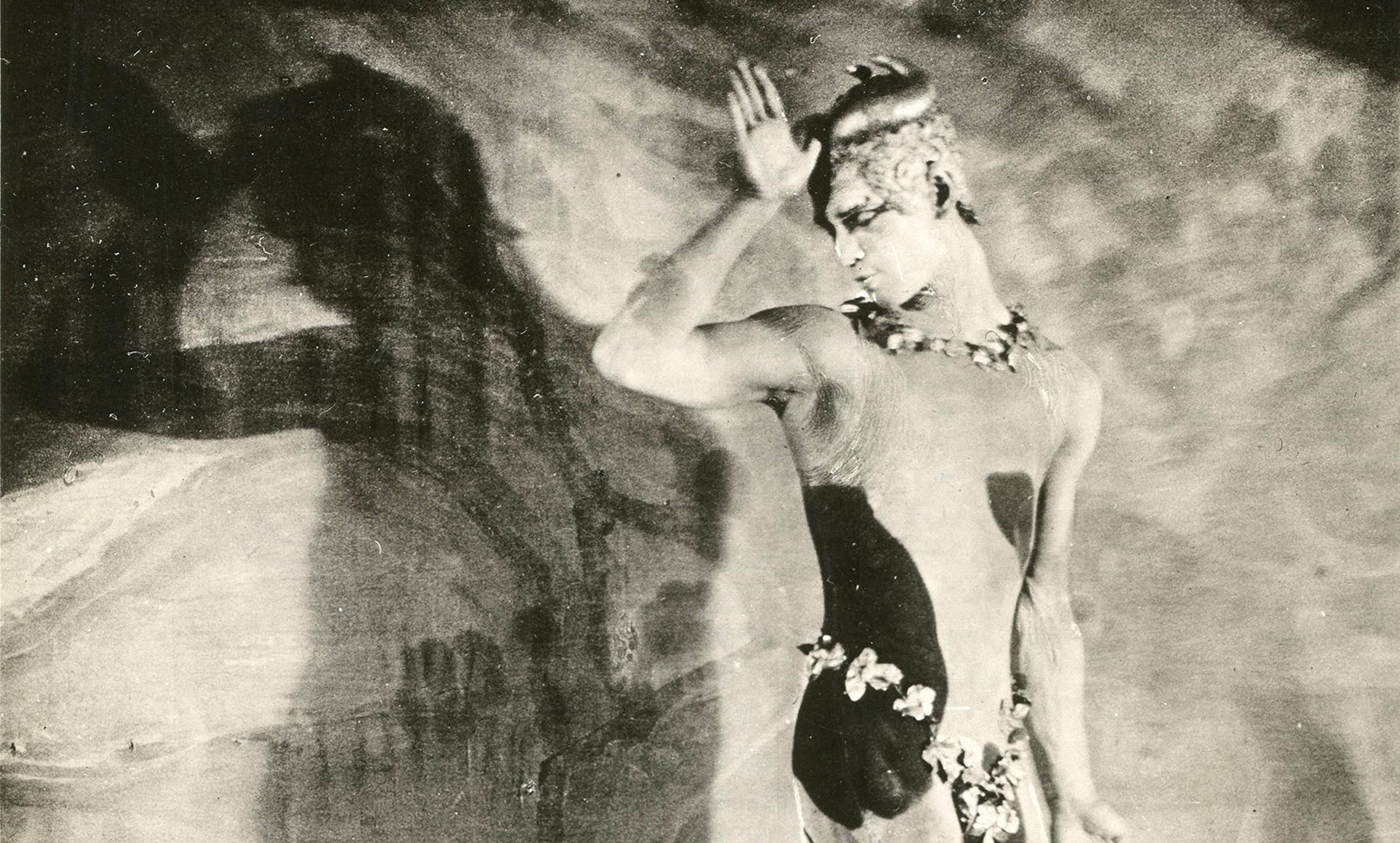 Black-and-white photo of a dancer in a costume performing against a shadowy background, with one hand raised near his head.