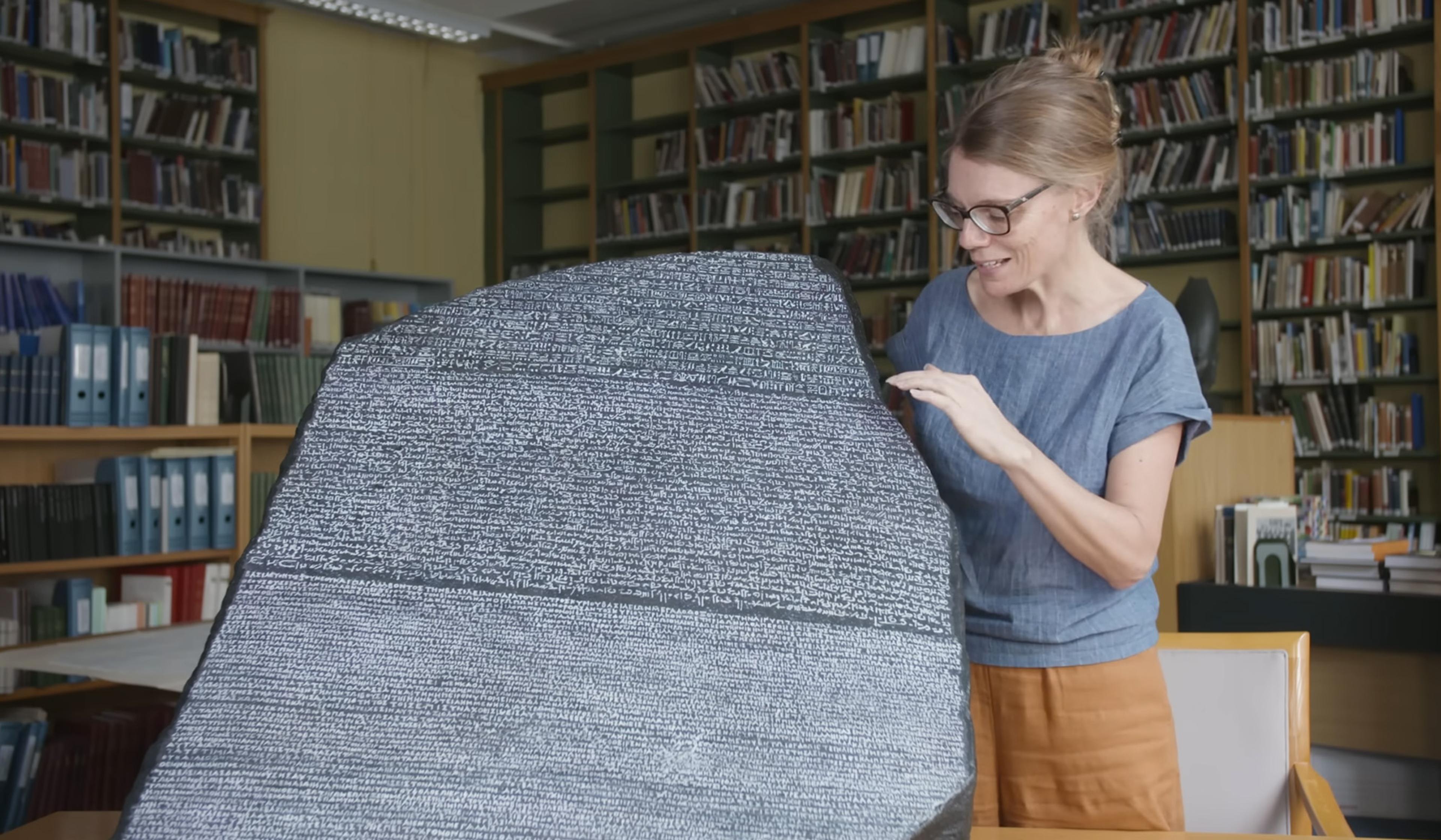 A woman examines the Rosetta Stone in a library setting filled with shelves of books and documents.