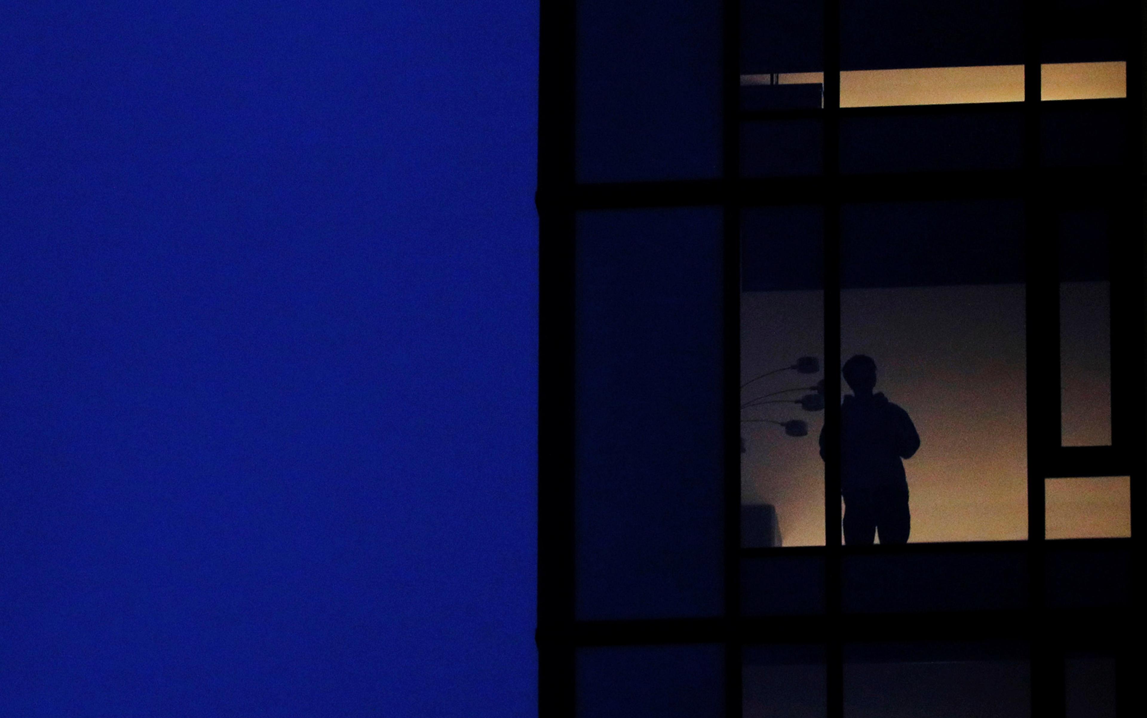 Silhouette of a person standing at a window inside a dimly lit building at night against a deep blue sky.