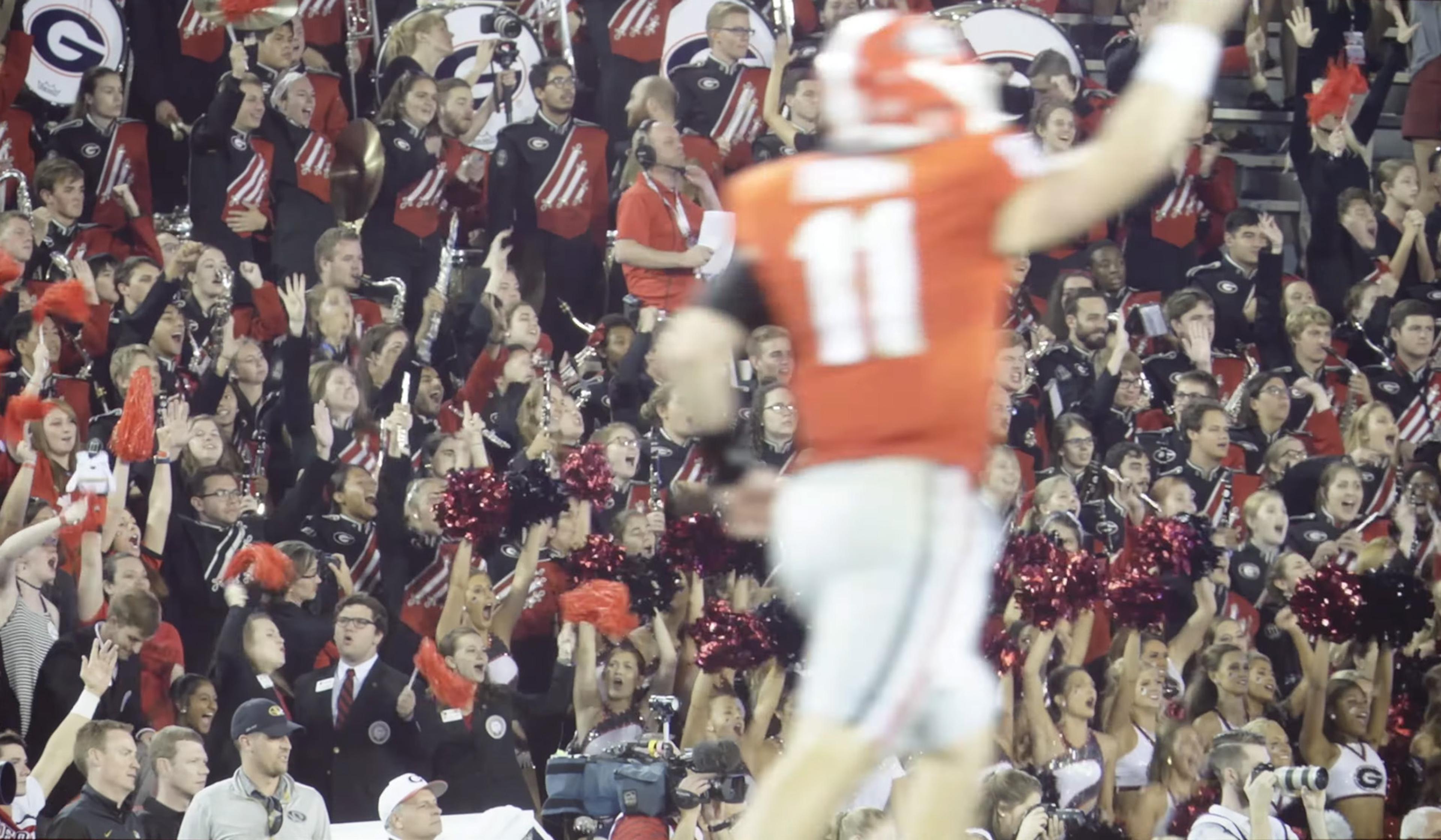 A football player is celebrating in front of an enthusiastic crowd, including a marching band and cheerleaders holding red pom-poms.