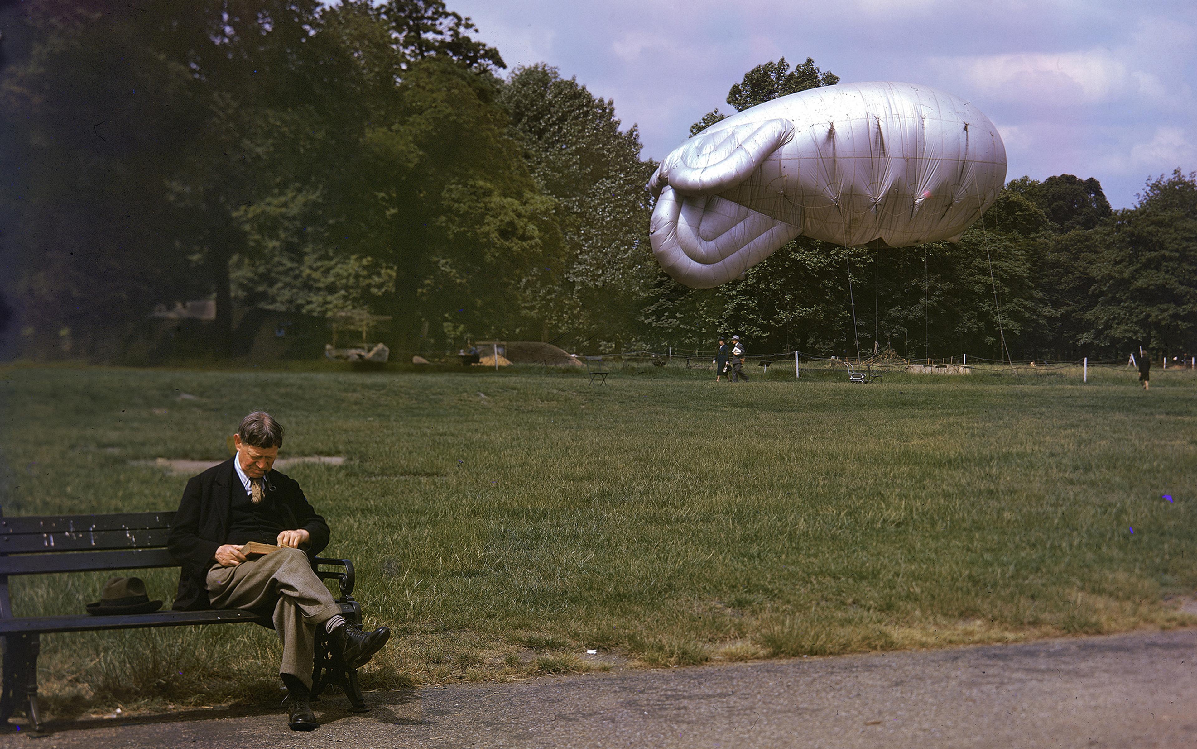 A man reading on a park bench with a silver blimp floating in the background among trees.