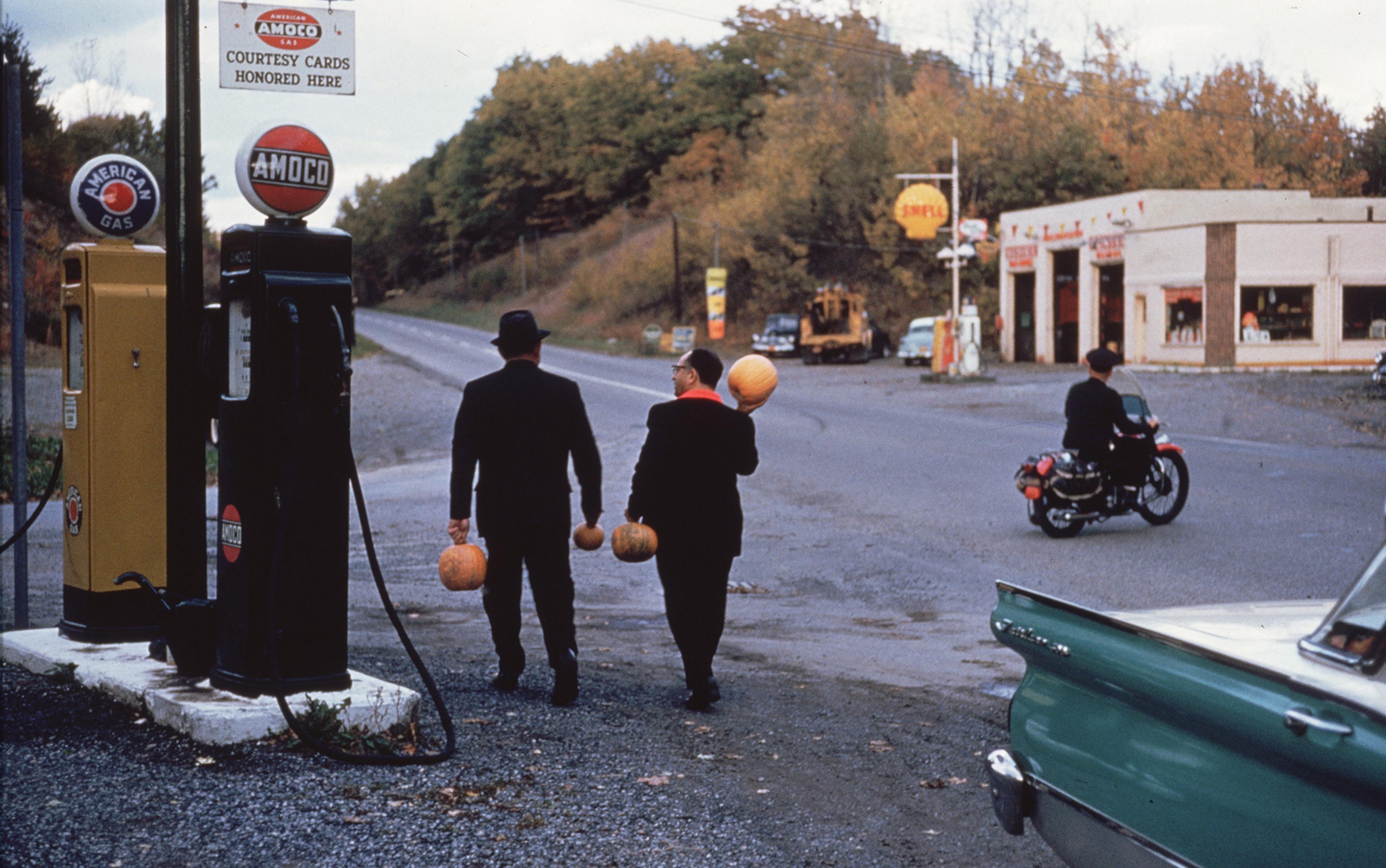 Two people carry pumpkins near an Amoco petrol pump with a motorcyclist and a garage visible in the background.