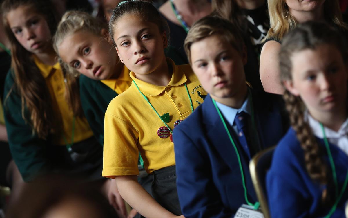 Children in uniforms sitting and listening attentively, with some wearing yellow and green while others are in blue blazers and ties.