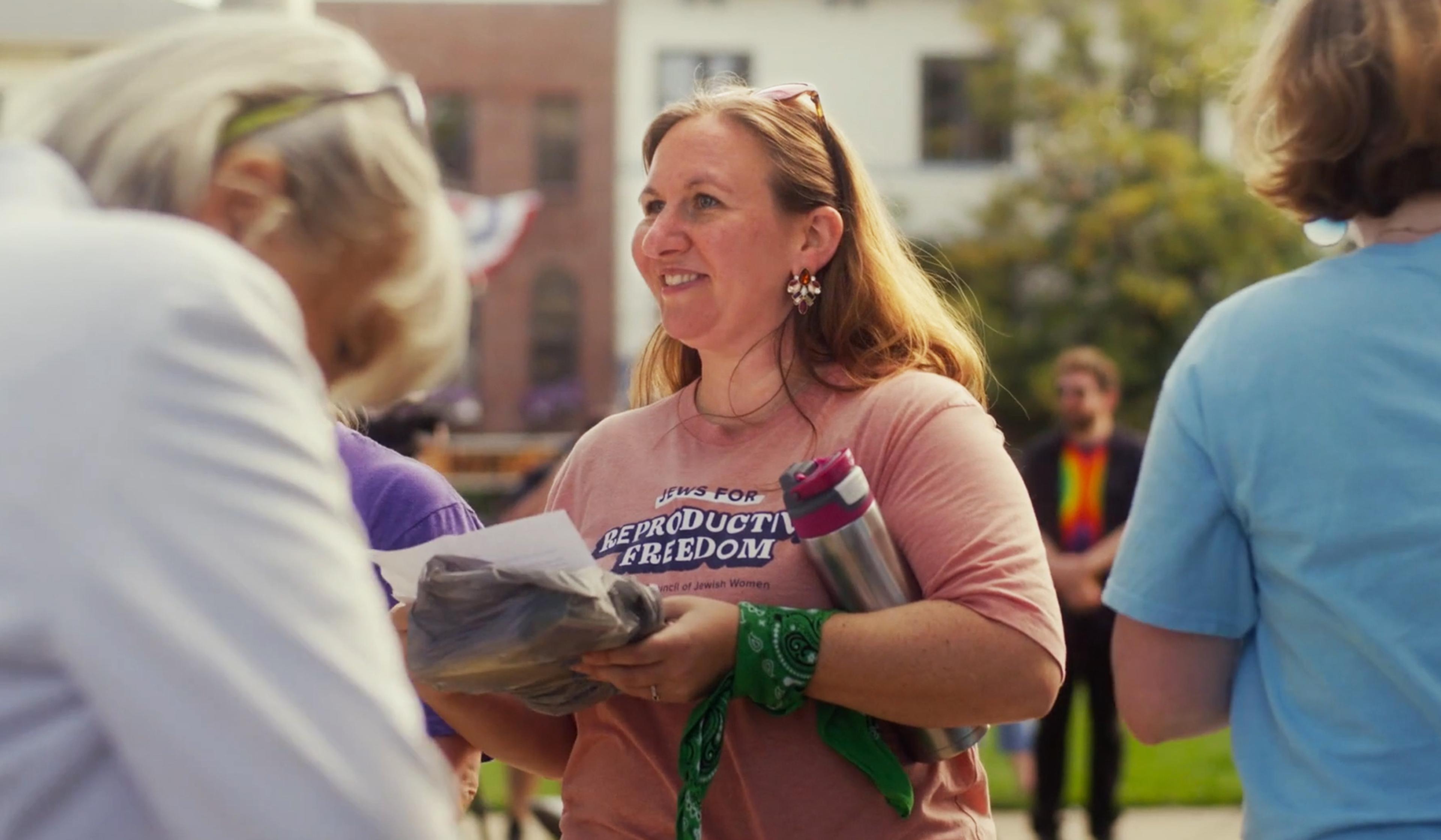 Woman with brown hair in a “Jews for Reproductive Freedom” shirt smiling and holding papers and a water bottle at an outdoor event.