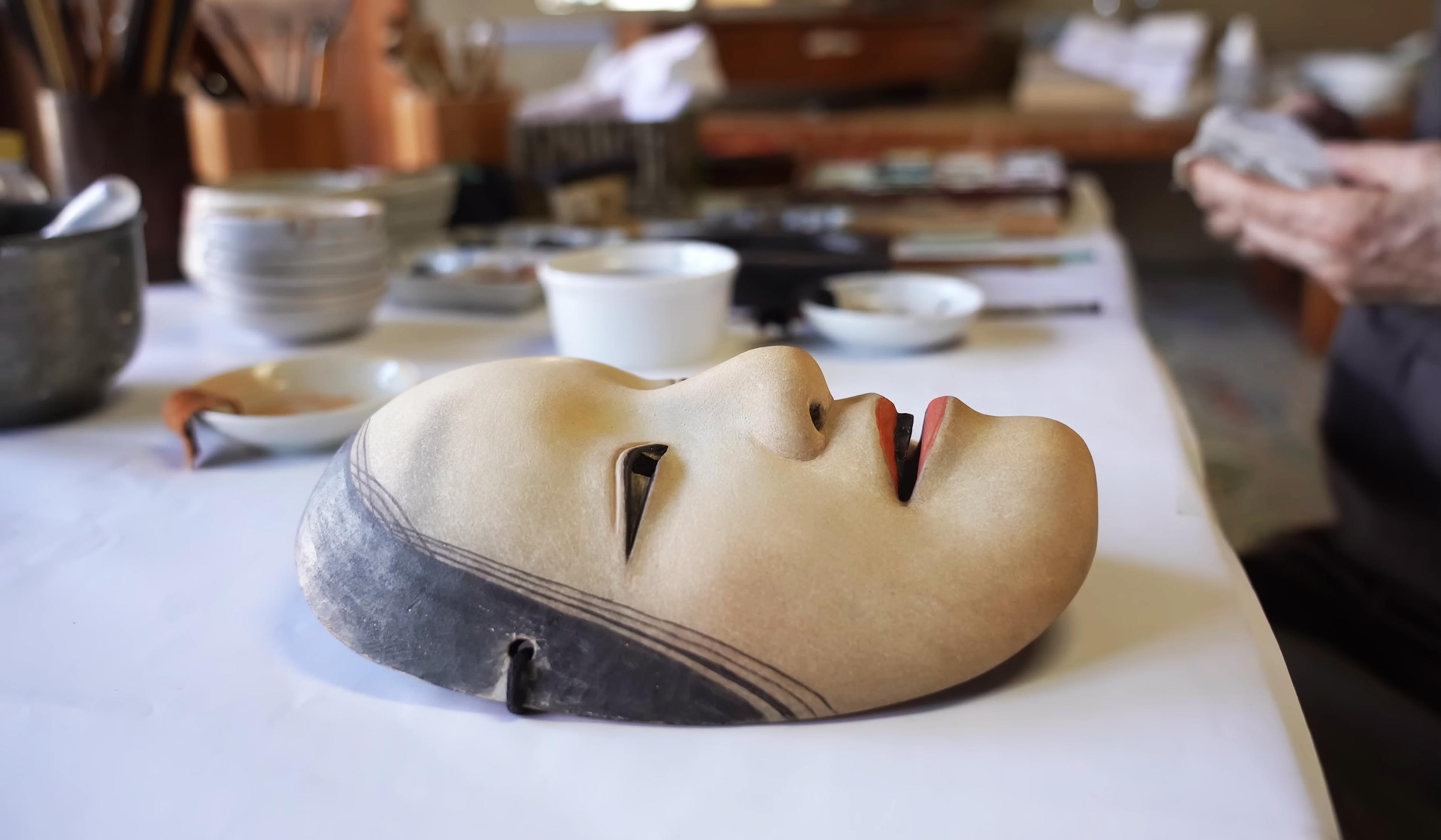 A traditional Noh theatre mask lies on a table, surrounded by pottery and art supplies in a workshop. A person works in the blurred background.