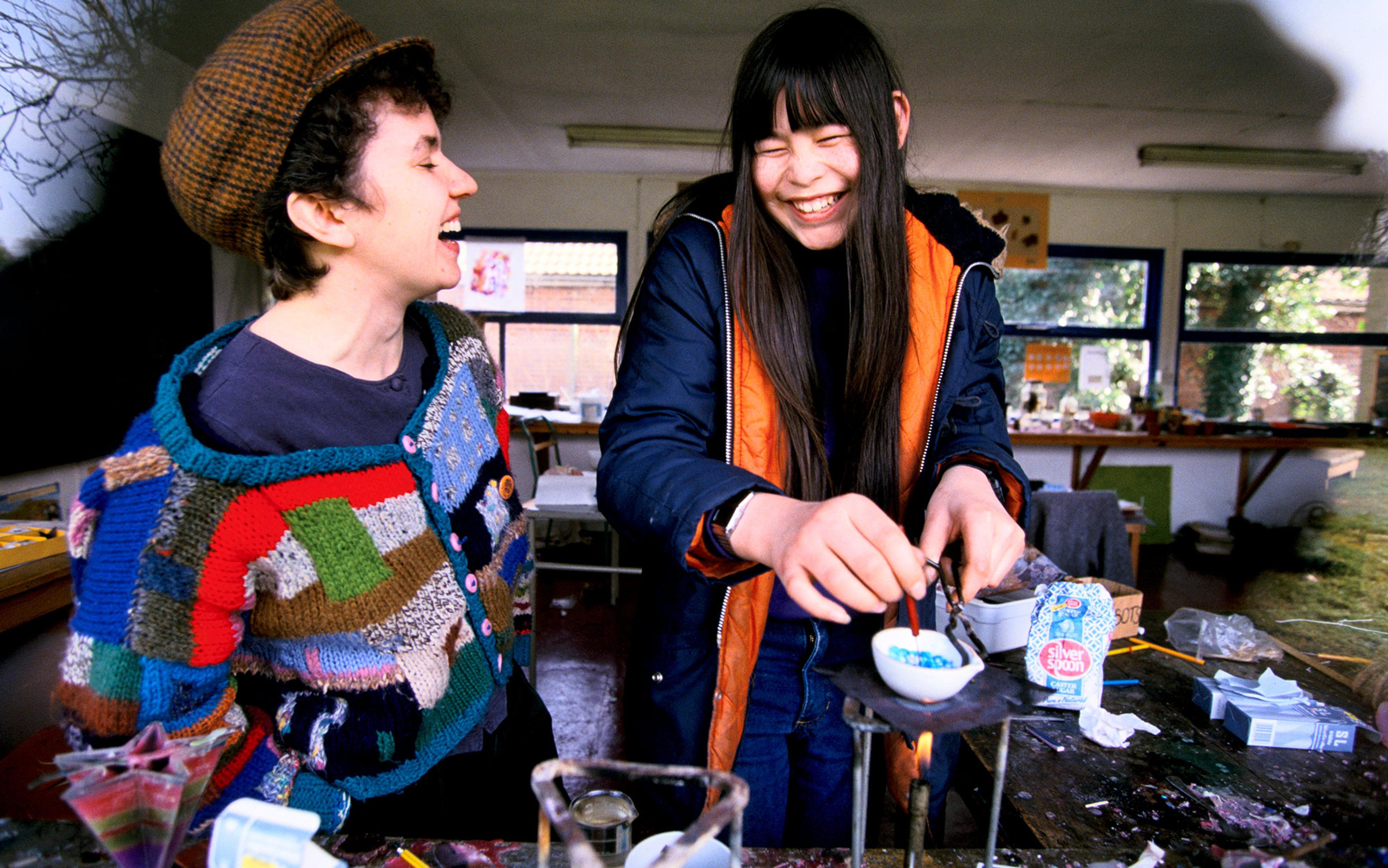 Two women laughing, one wearing a patchwork jumper and hat, the other melting materials in a bowl over a flame in a workshop.