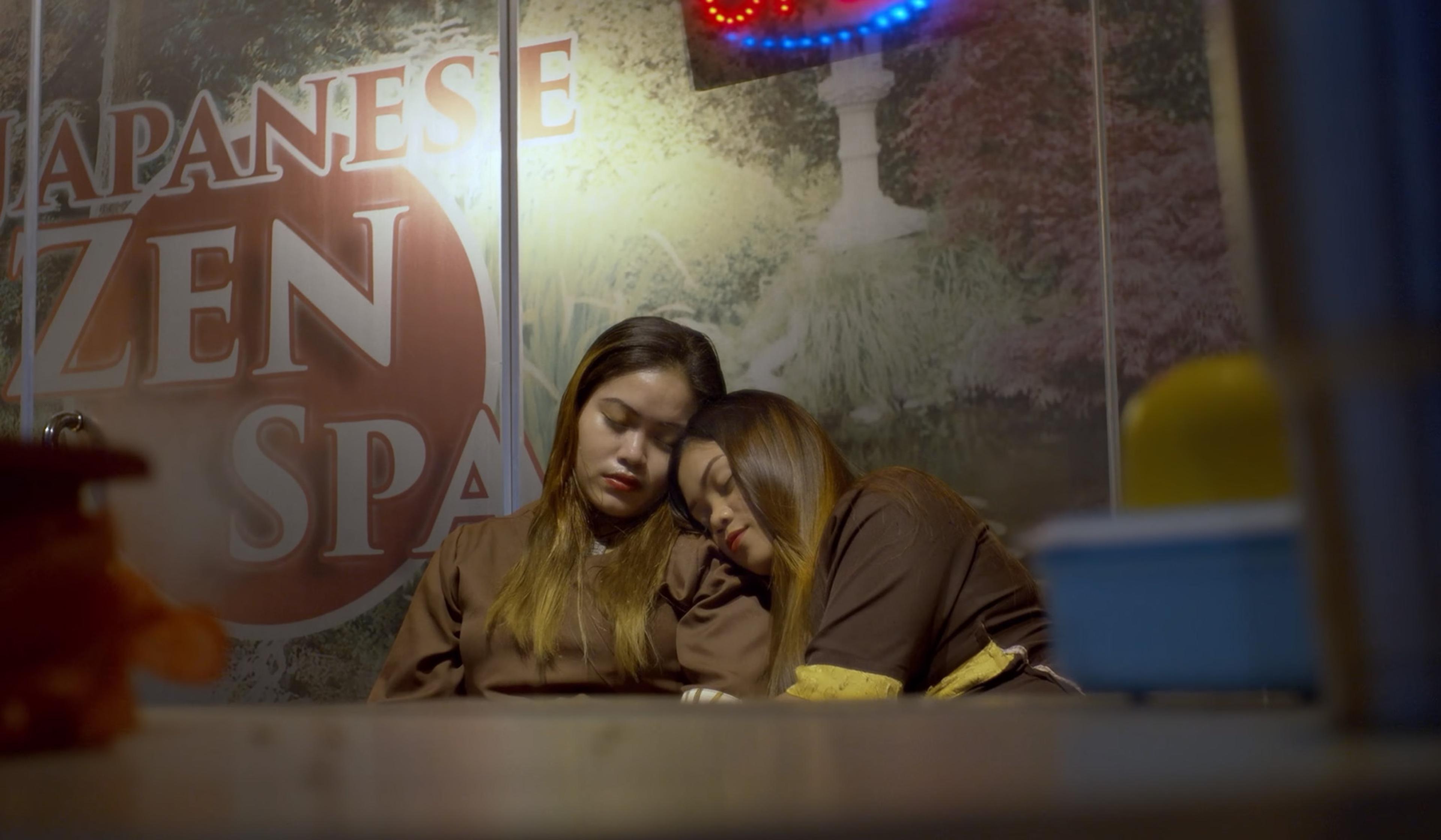 Two women sleeping and leaning on each against a large sign reading ‘Japanese Zen Spa’.