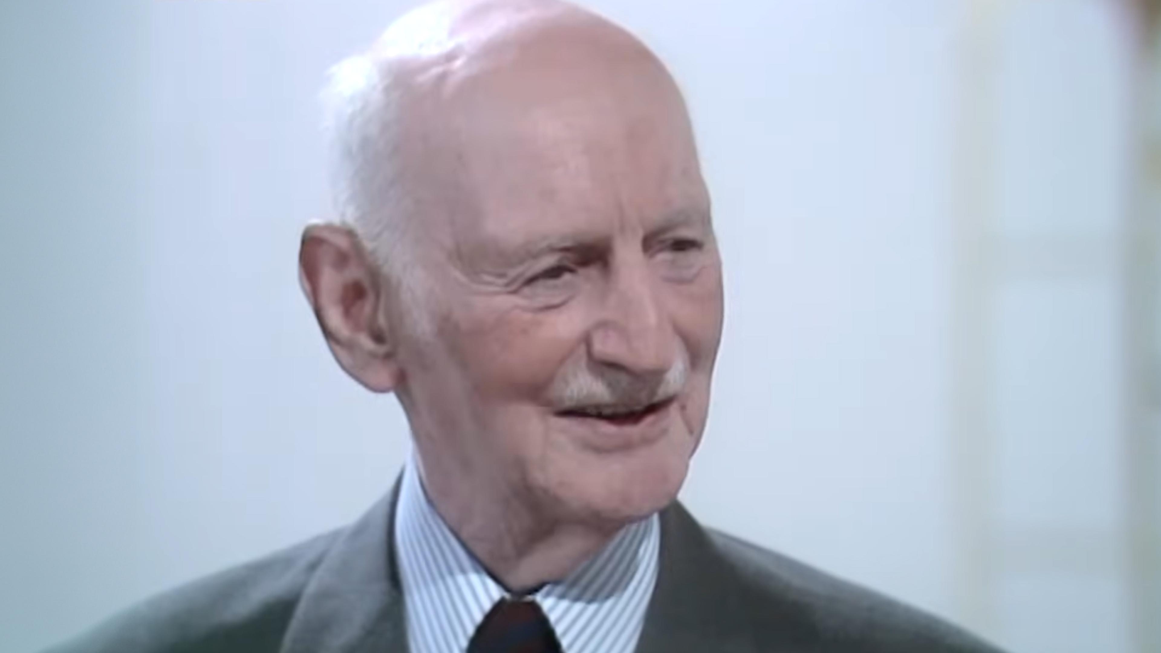 Photo of an elderly man with a bald head and moustache wearing a suit and tie, smiling against a light-coloured, blurred background.