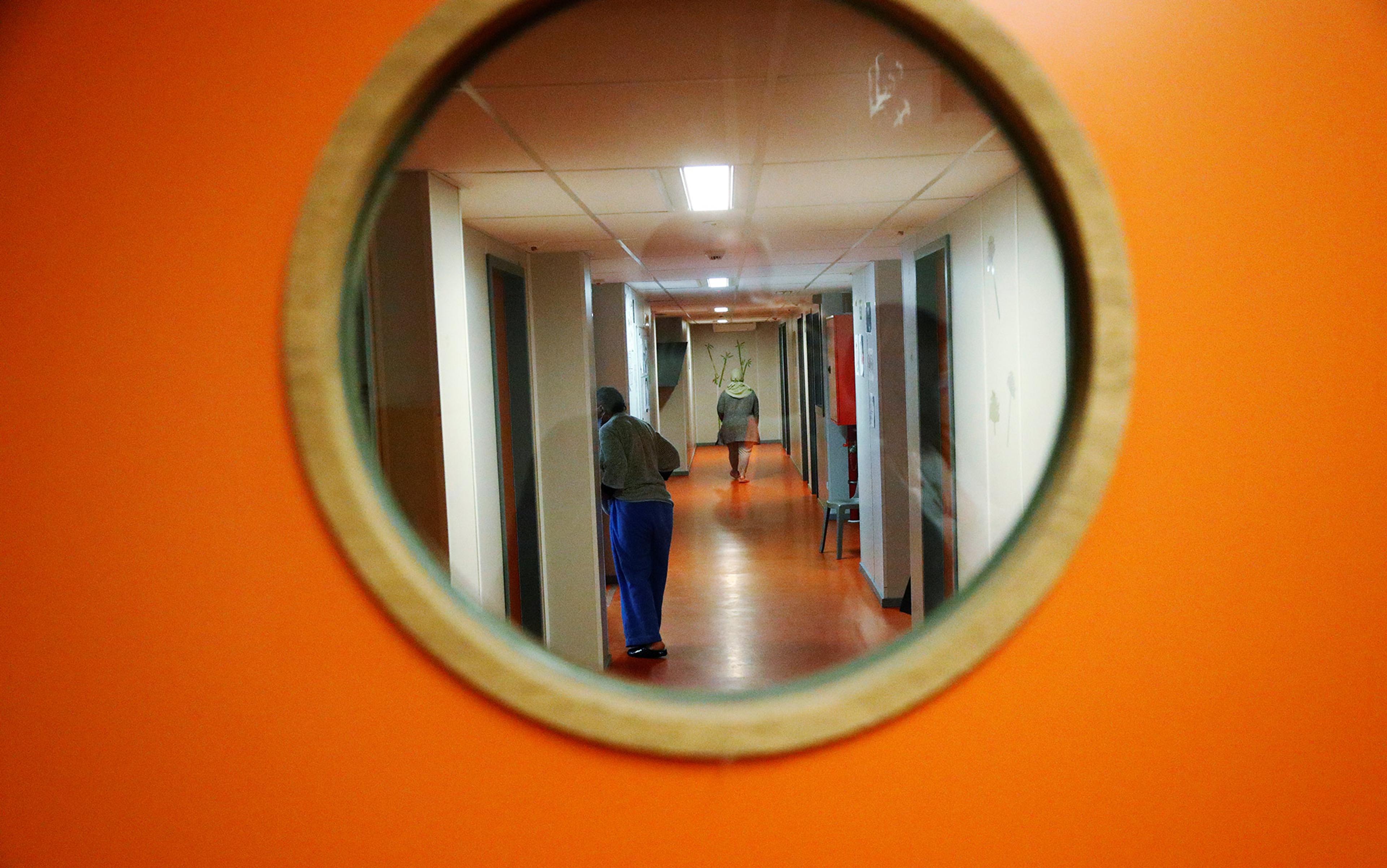 View through a round window on an orange door, showing two people walking down a brightly lit corridor with orange floors and white walls.