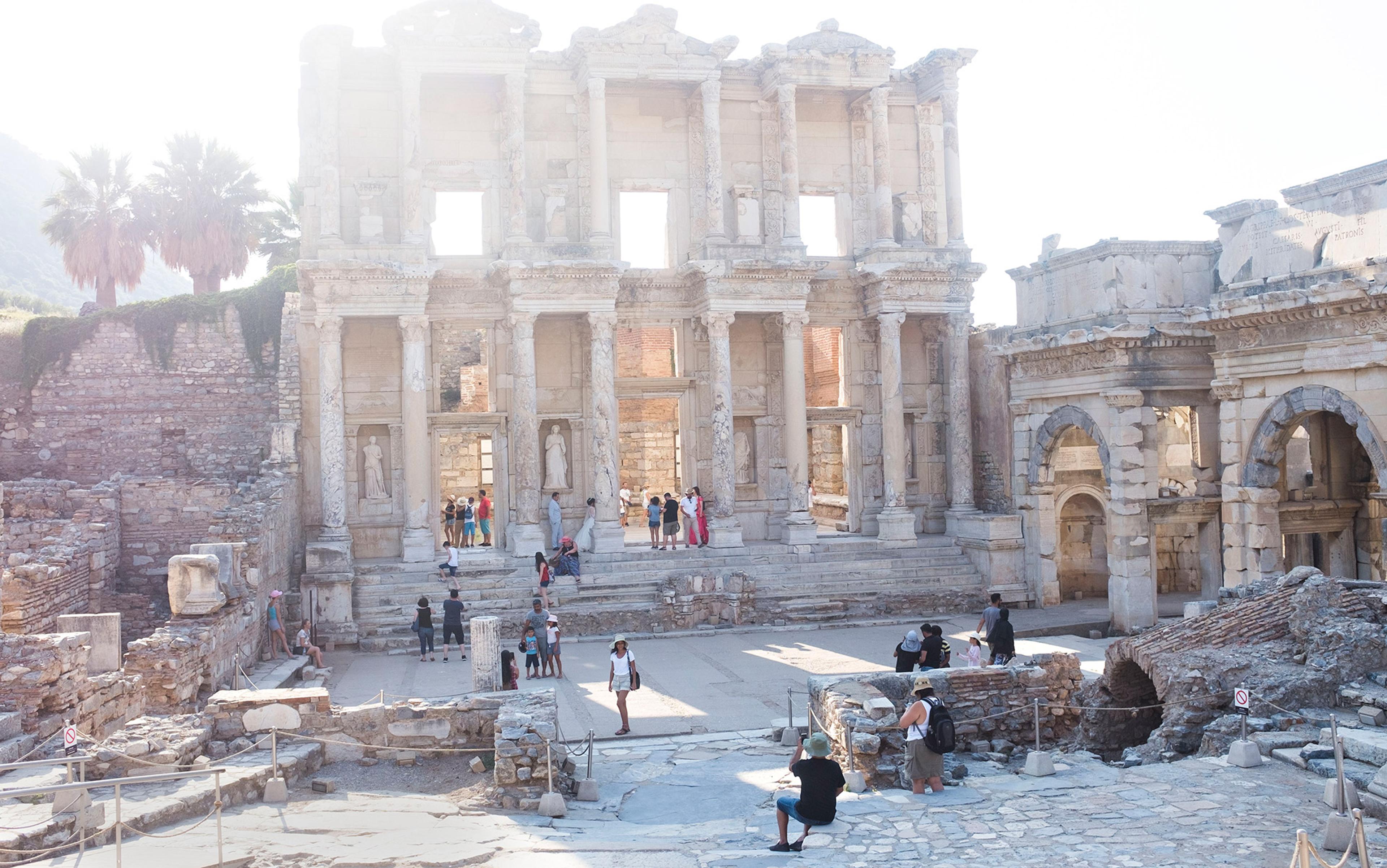 Ancient ruins with arches and columns, tourists exploring, and palm trees in the background, in a bright, sunny setting.