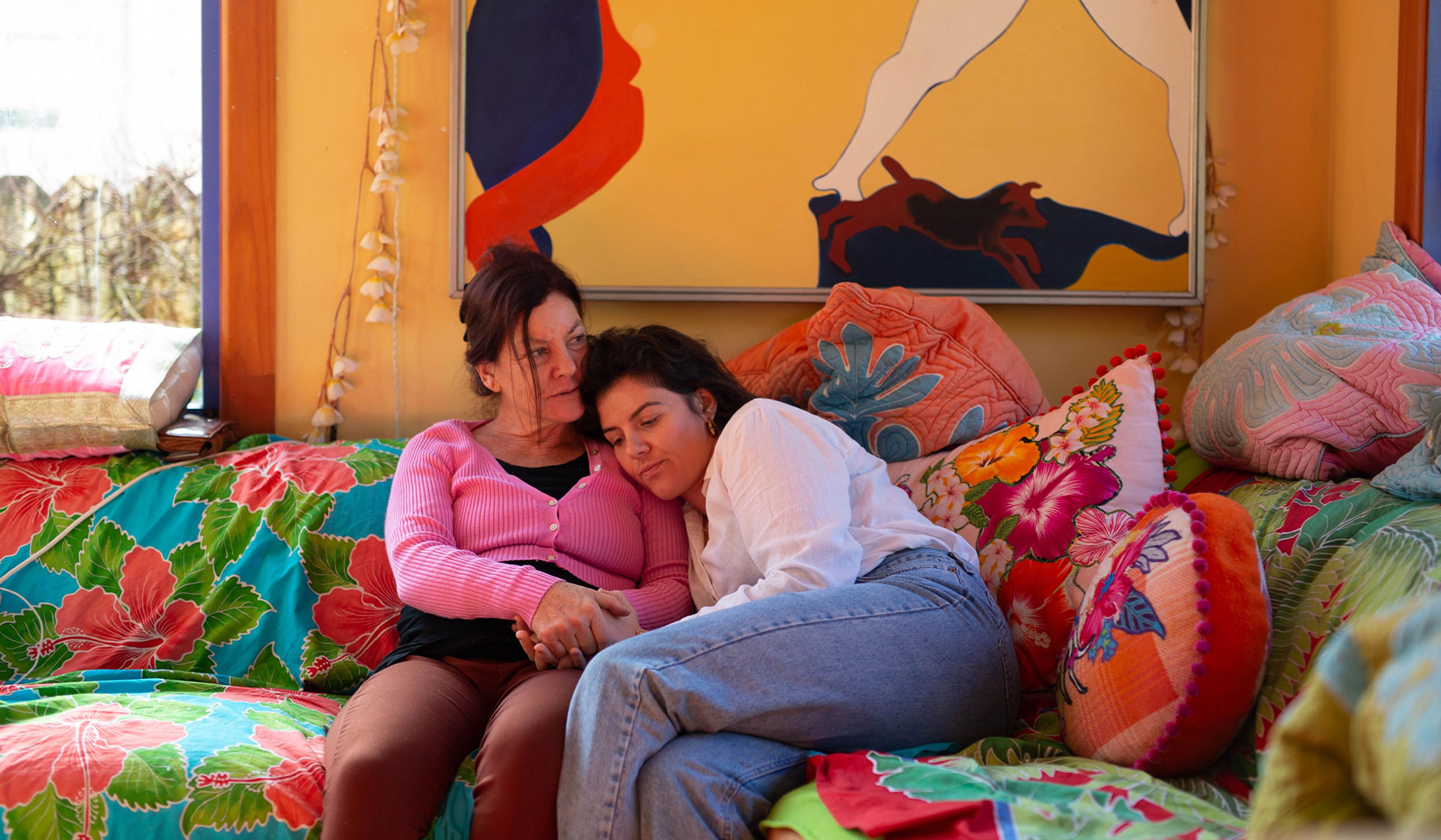 A young woman leans against an older woman on a sofa with floral cushions and a painting on the wall behind.