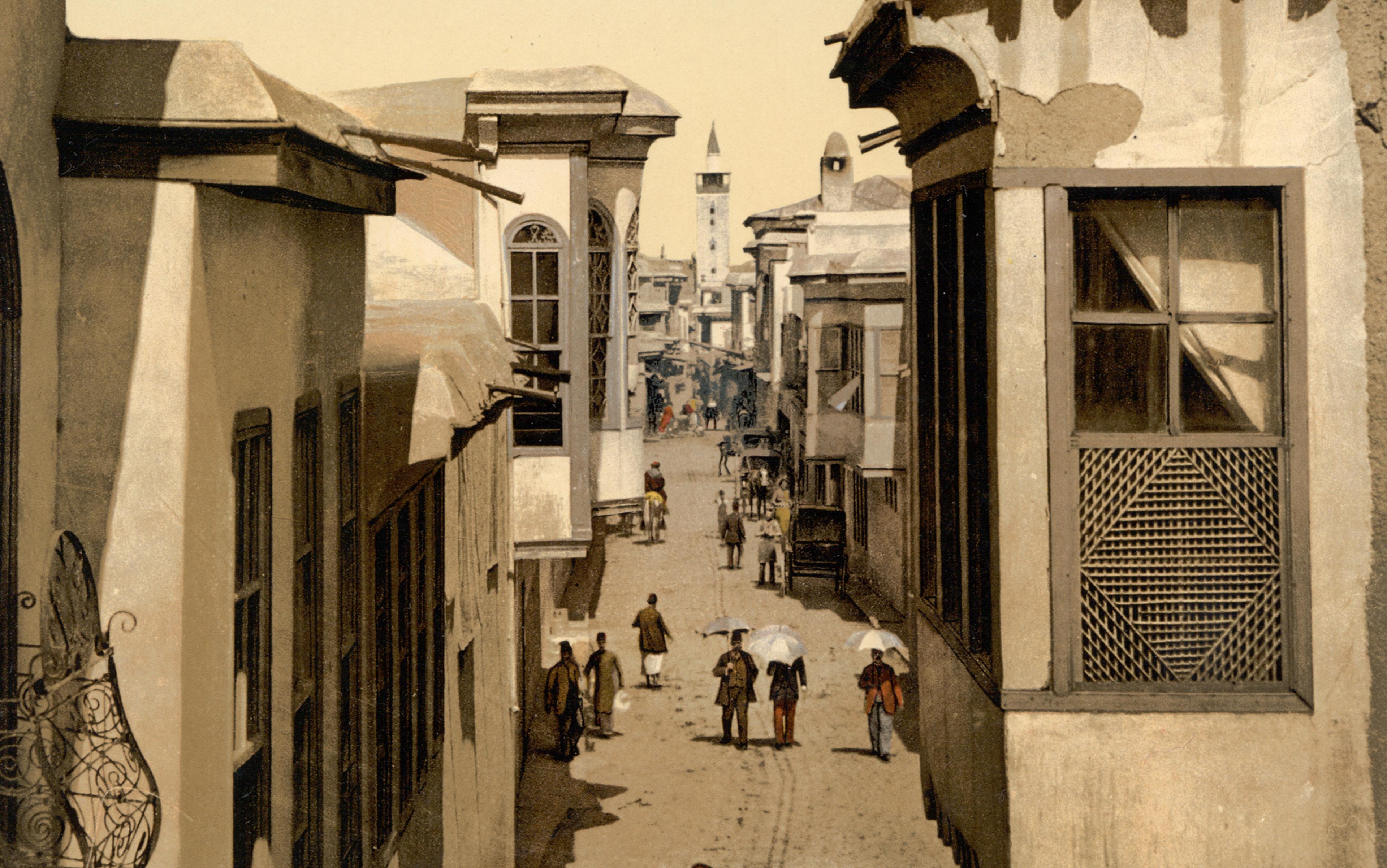 Photochrom image of a narrow street lined with Middle-Eastern buildings; people are walking down the middle of the street and some are holding umbrellas.