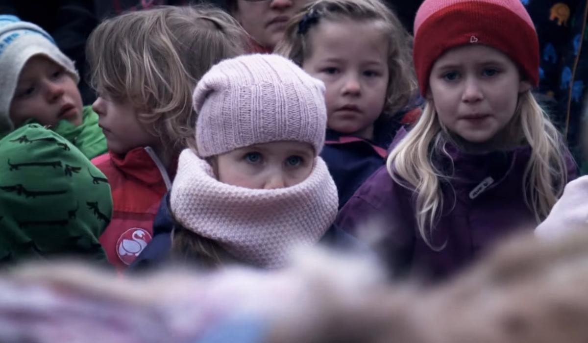 Group of children dressed in winter clothing, including hats and scarves, standing outside and looking forward with solemn expressions.