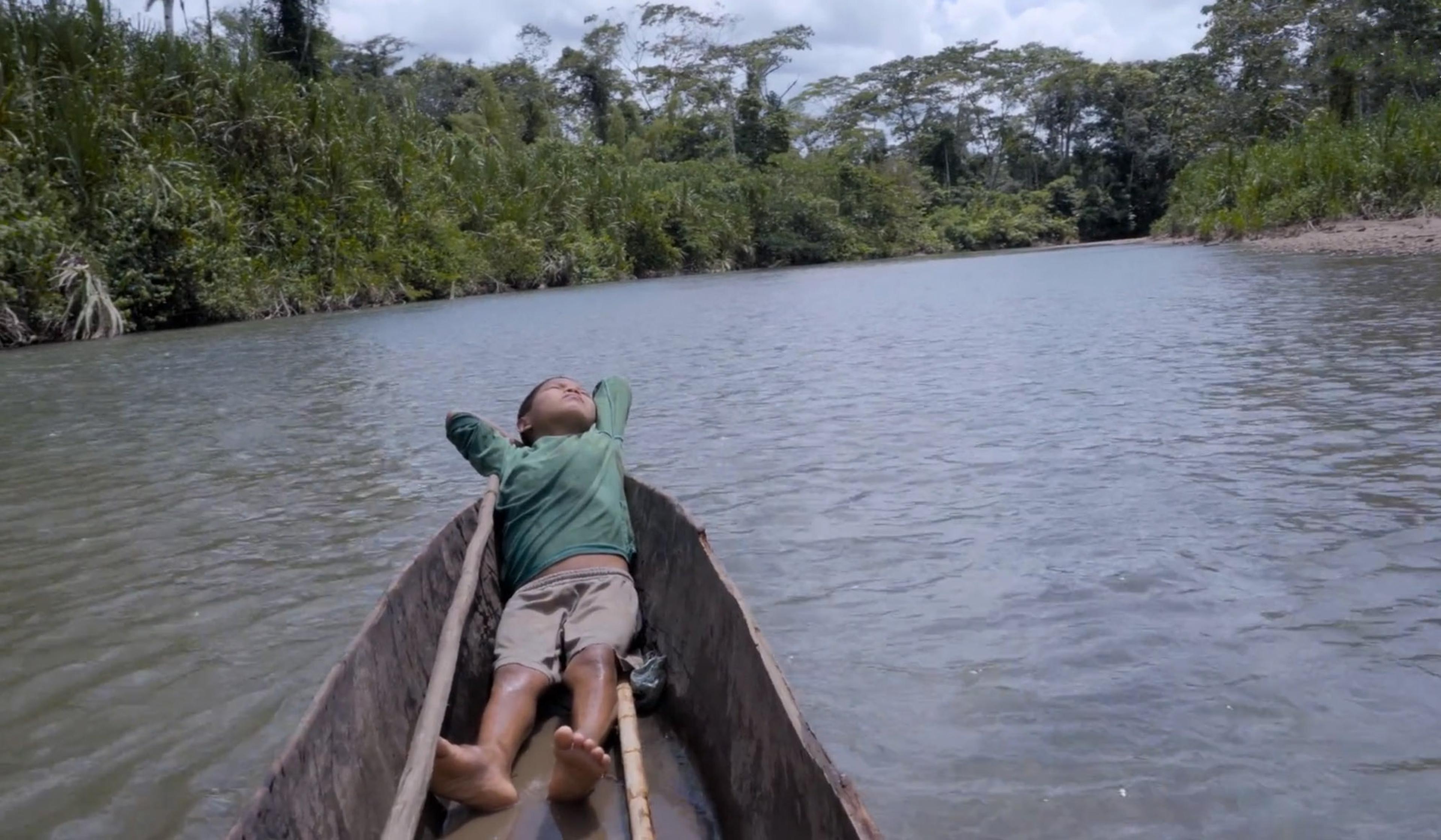 A child in a green shirt and shorts lying in a wooden canoe on the Ecuadorian Amazon river surrounded by dense forest.