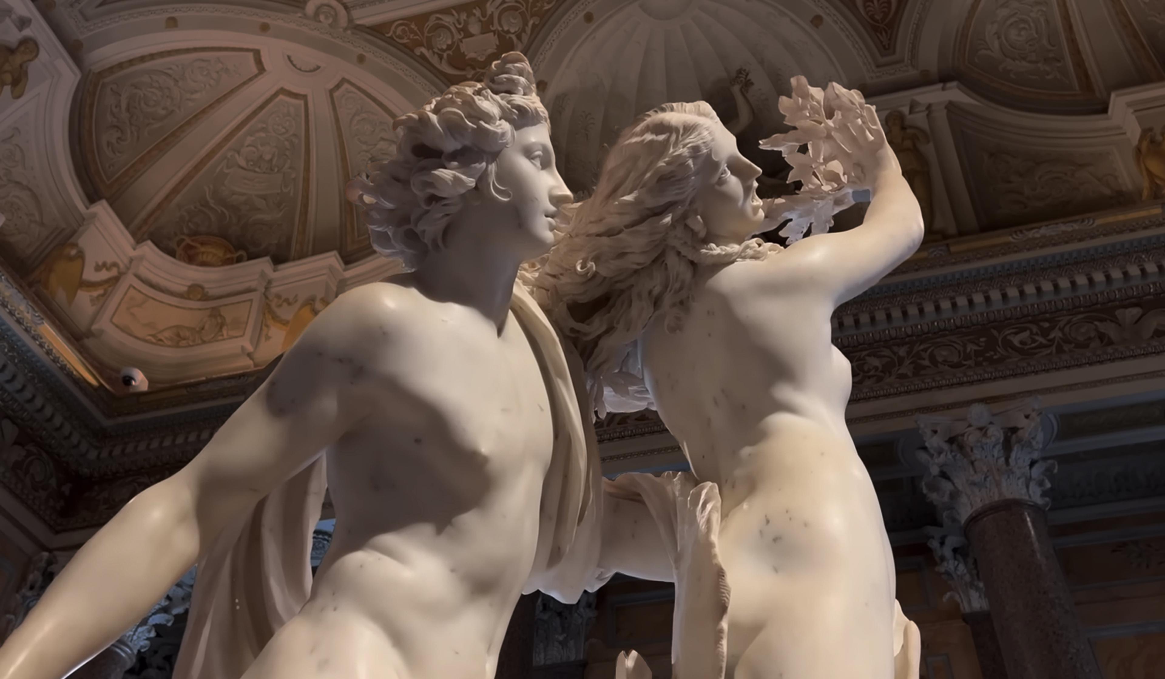 Marble statue depicting Apollo and Daphne, set against a detailed, ornate ceiling in a classical style room. Daphne is reaching upward with flowing hair.