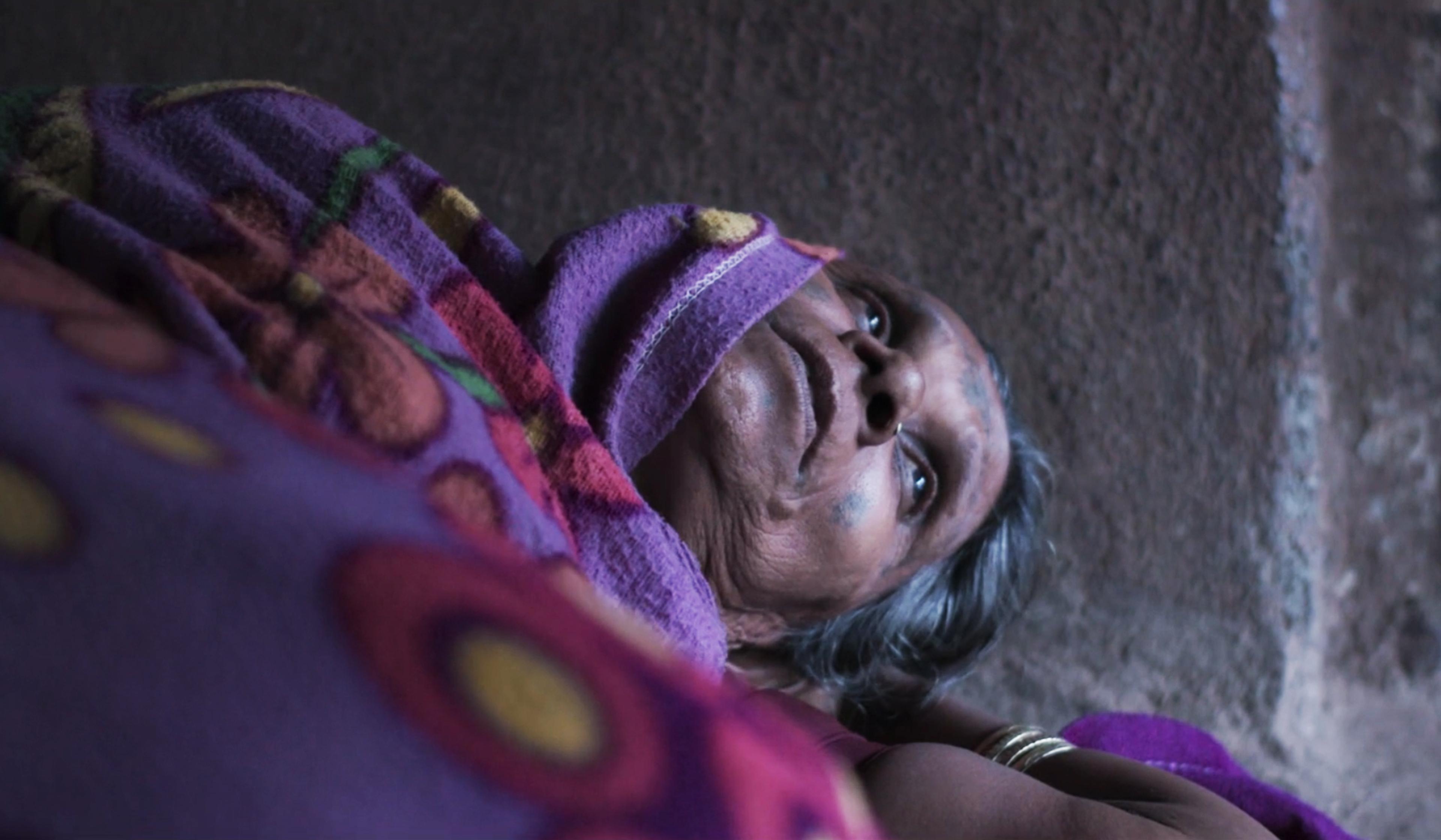 A woman with grey hair lies down, partially covered with a colourful, patterned blanket against a textured wall.