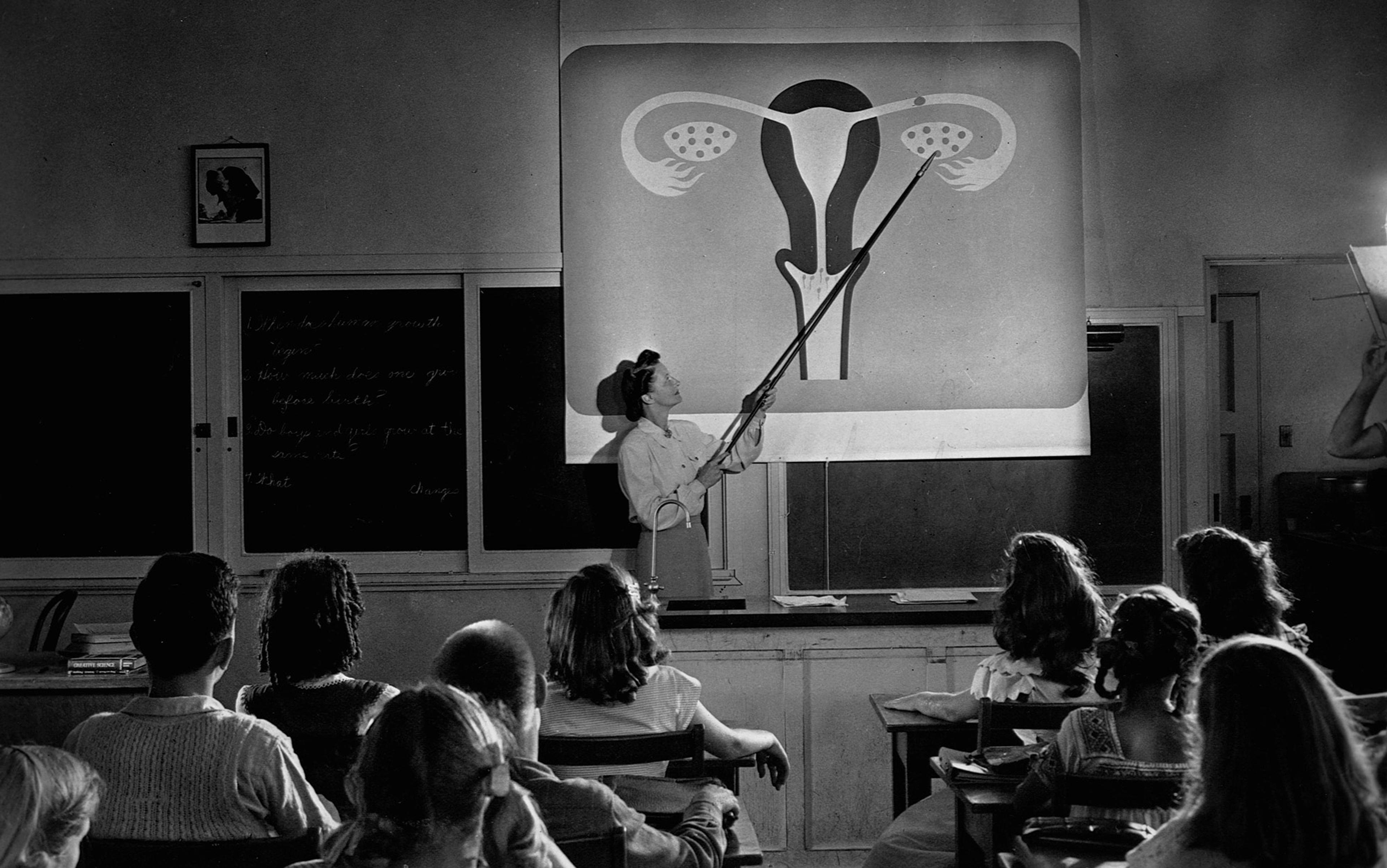 Black-and-white photo of a teacher pointing to a diagram of the female reproductive system, students watching attentively.