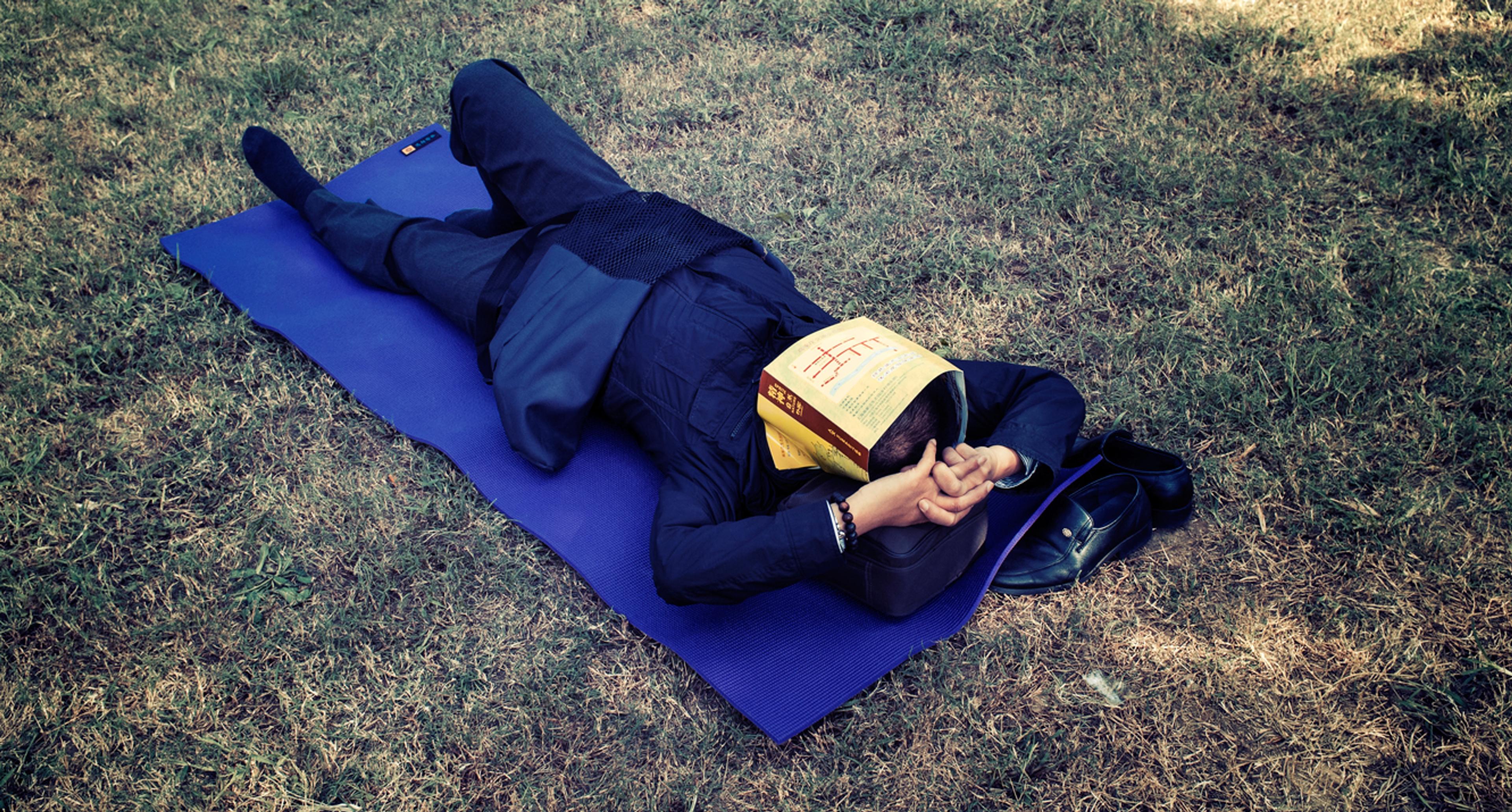 Photo of a person lying on a blue mat on grass with a book covering their face, hands clasped, shoes beside them.