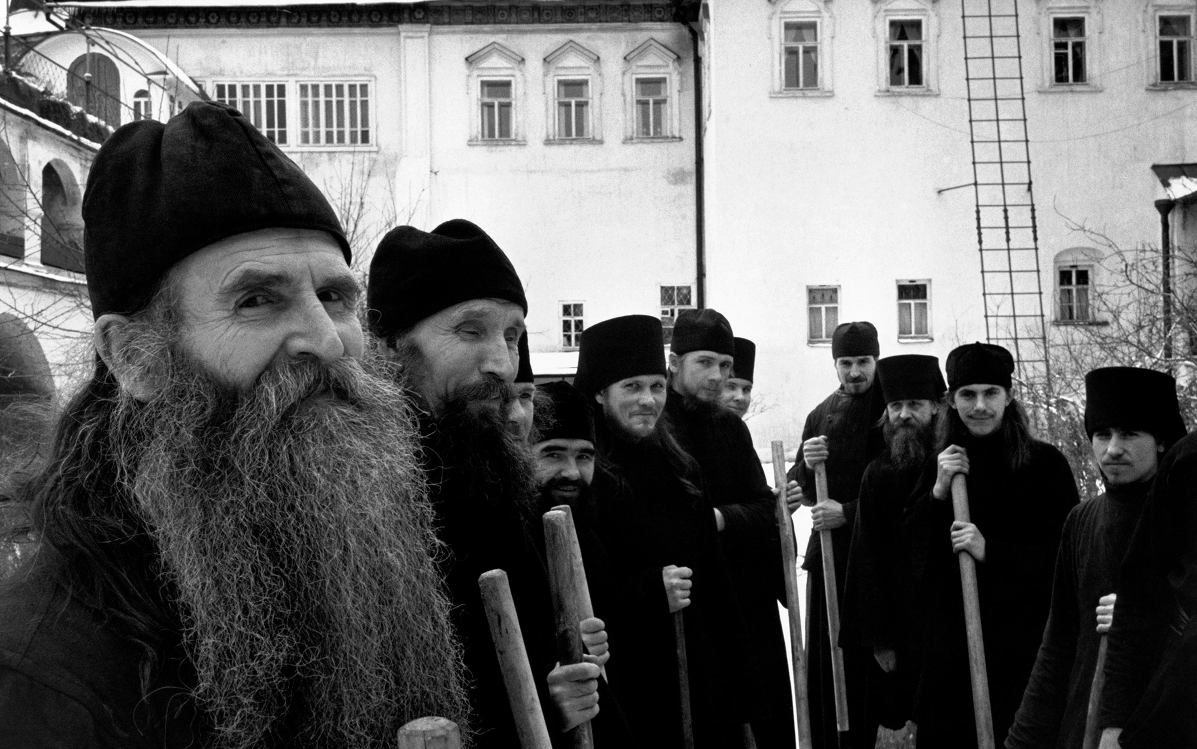 A group of bearded men wearing dark clothing and hats, holding long wooden sticks, standing in front of a white building with arched windows.