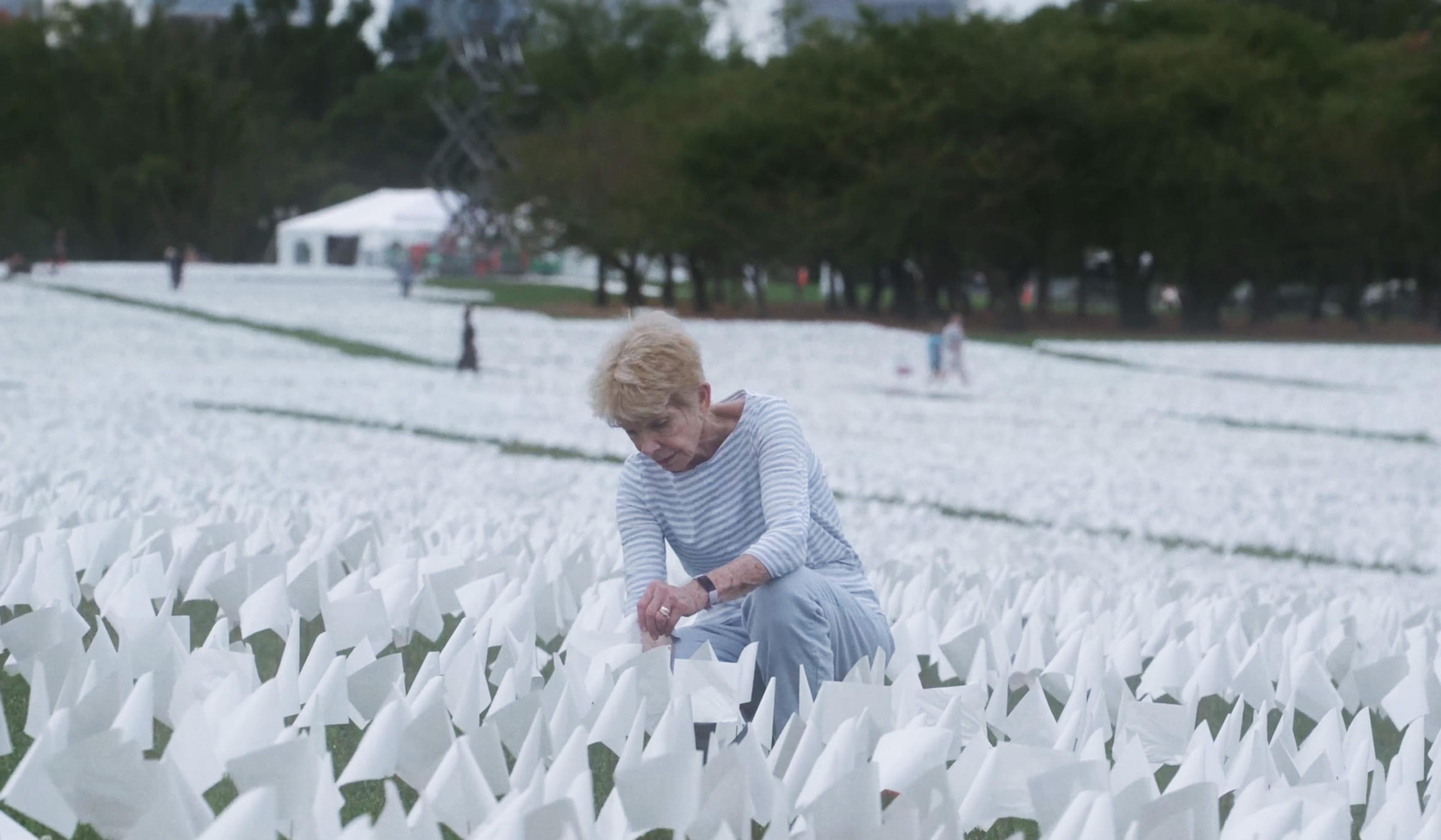 A woman kneels among thousands of white flags in a grassy field, with a white tent and trees in the background.