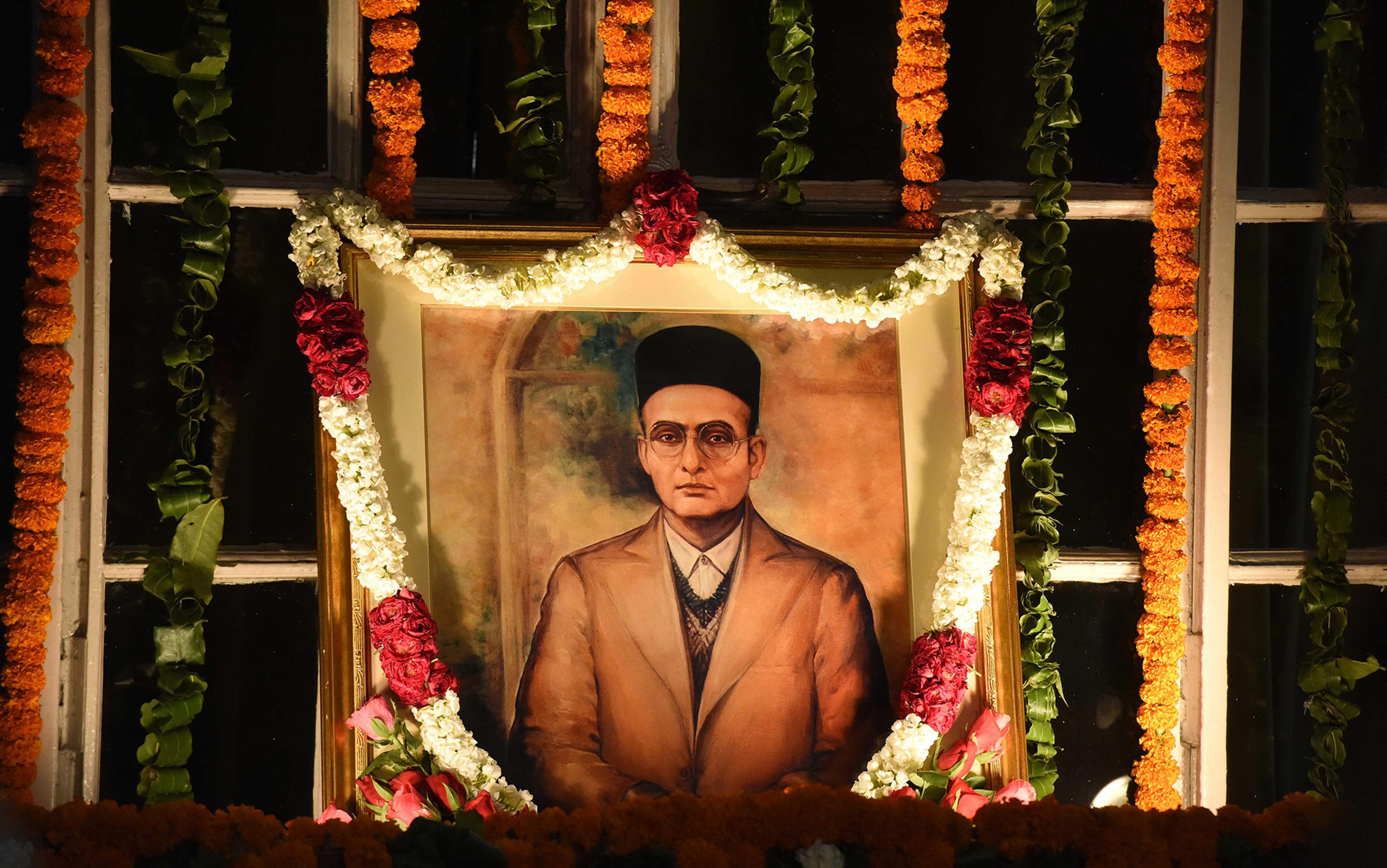 A portrait of a man with glasses and a cap, adorned with garlands of flowers, and surrounded by strings of marigolds and leaves.