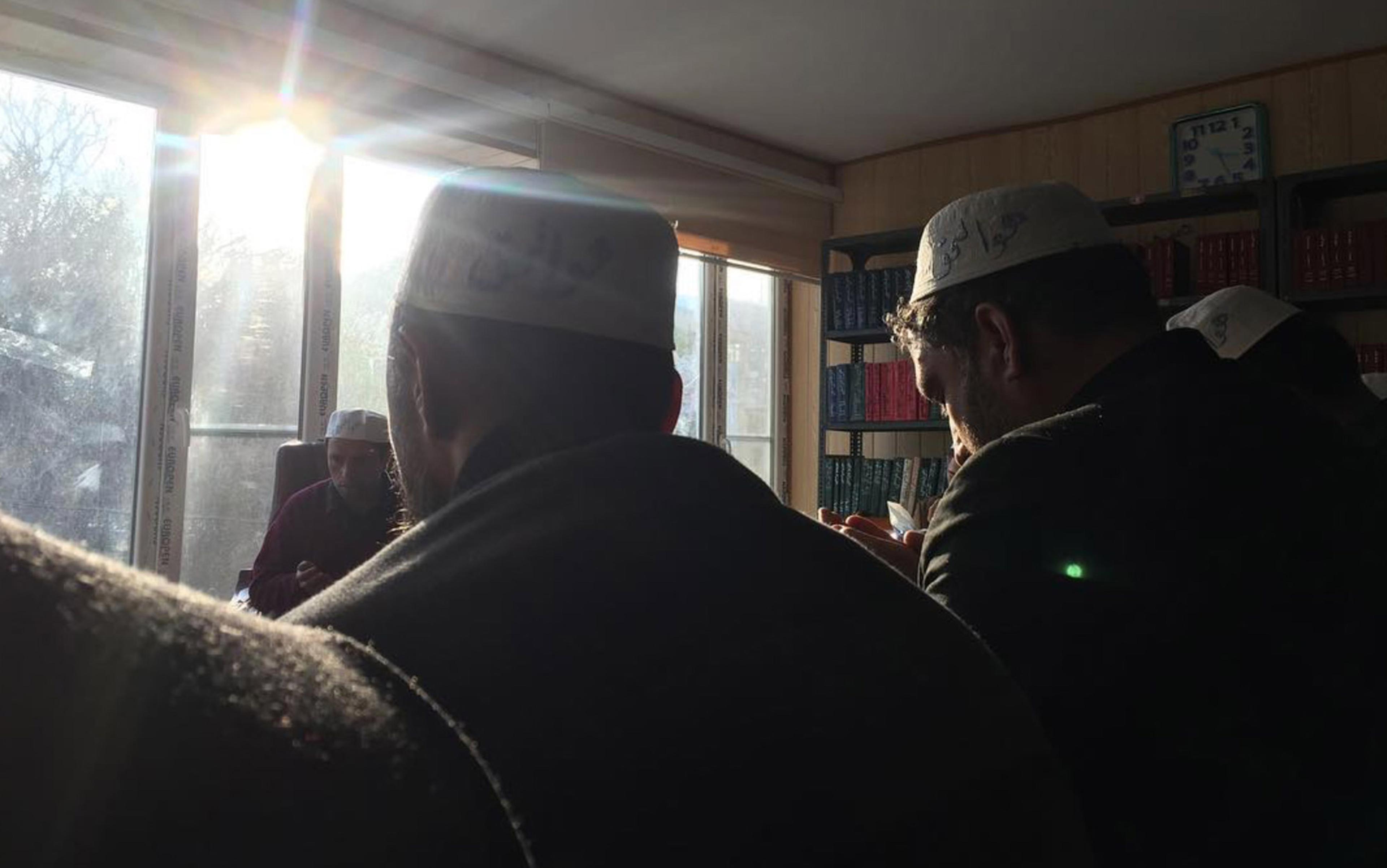 Men are seen in a nondescript office type rooma against sunlight streaming through the window, their hands raised in devotion