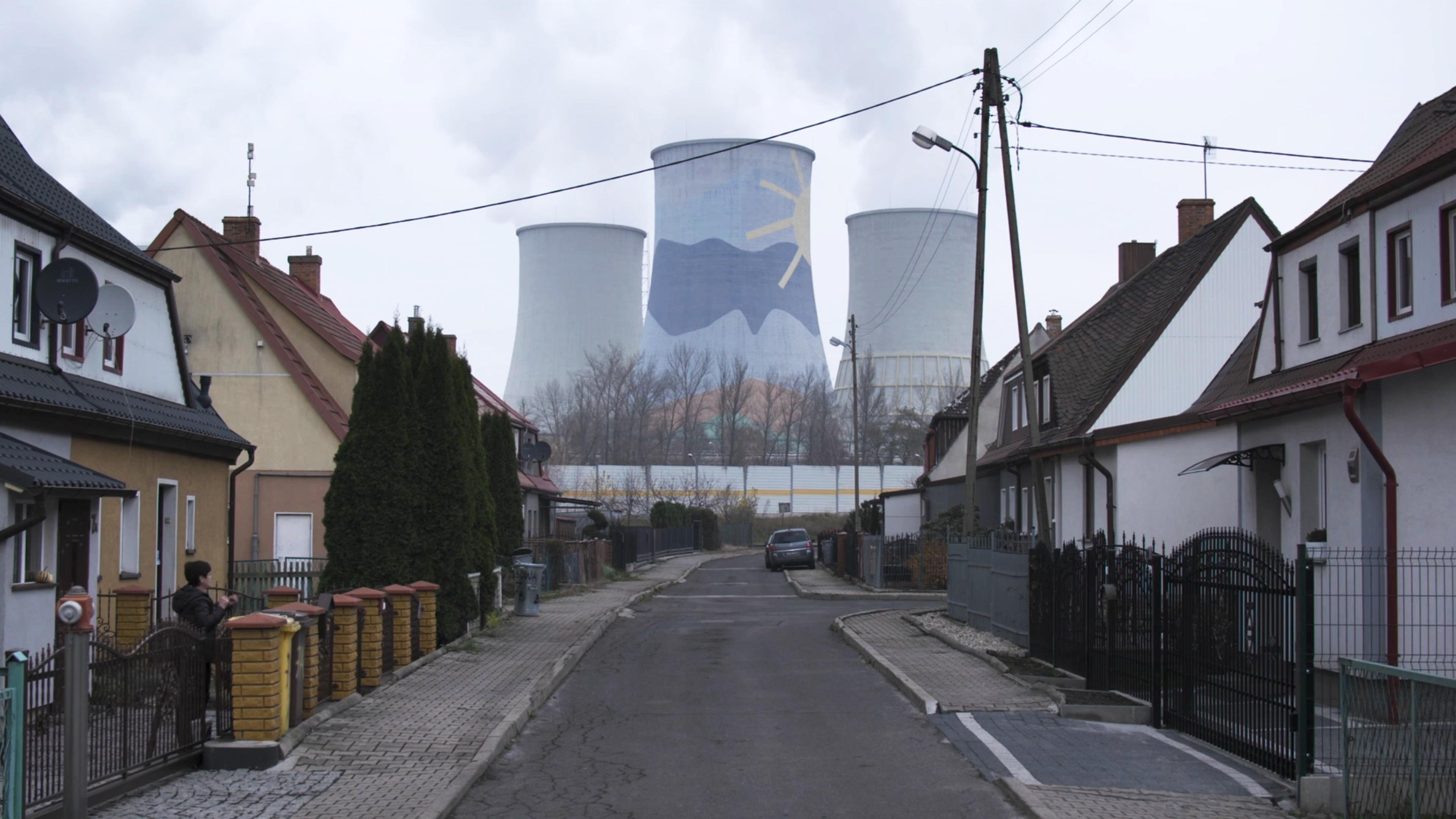 Residential street with terraced houses and power plant cooling towers in the background, one of which has a mural depicting a sun and mountains.