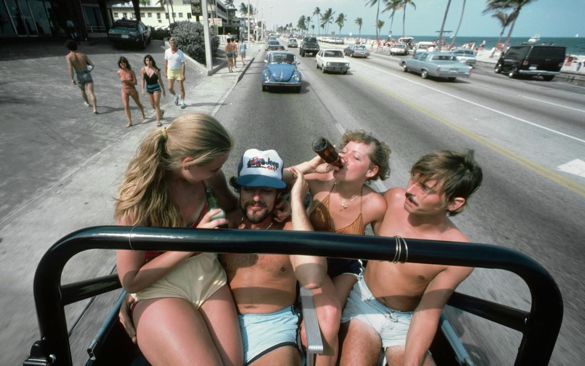 Two men and two women in beachwear sit in an open-top car on a palm-lined beach road. Two of them drink from bottles.