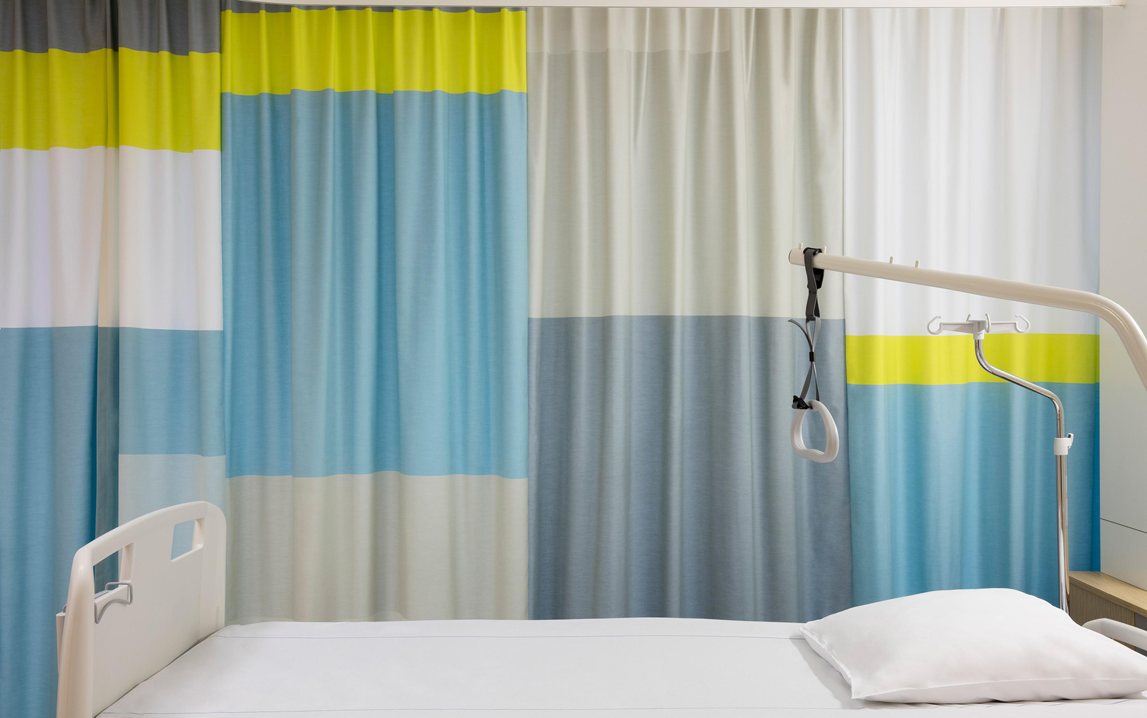 Hospital bed with colourful curtain background, white pillow, overhead patient lift, and hand control.