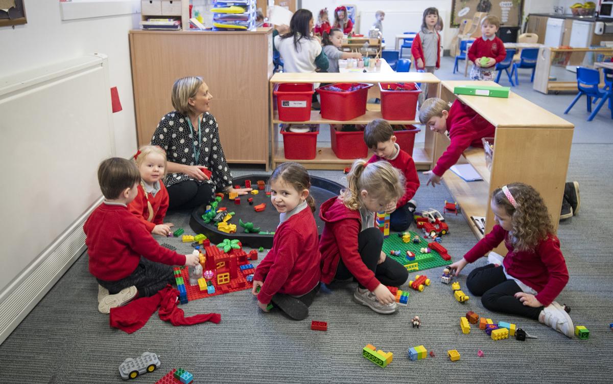 A group of young children and a teacher play with colourful building blocks in a classroom, with shelves and other students in the background.