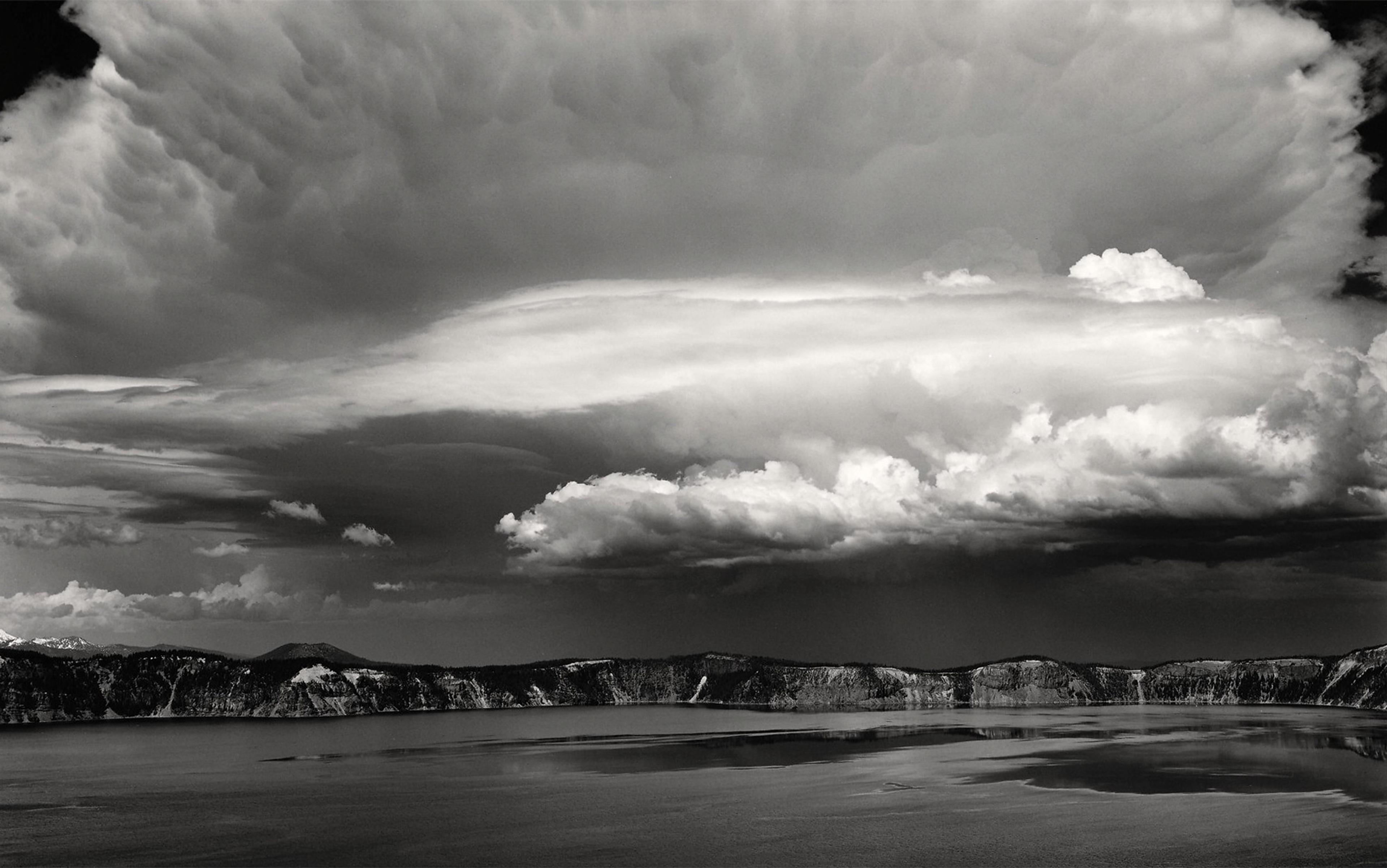 A dramatic black-and-white landscape of a vast lake, surrounded by mountains, under a large, turbulent sky filled with clouds.